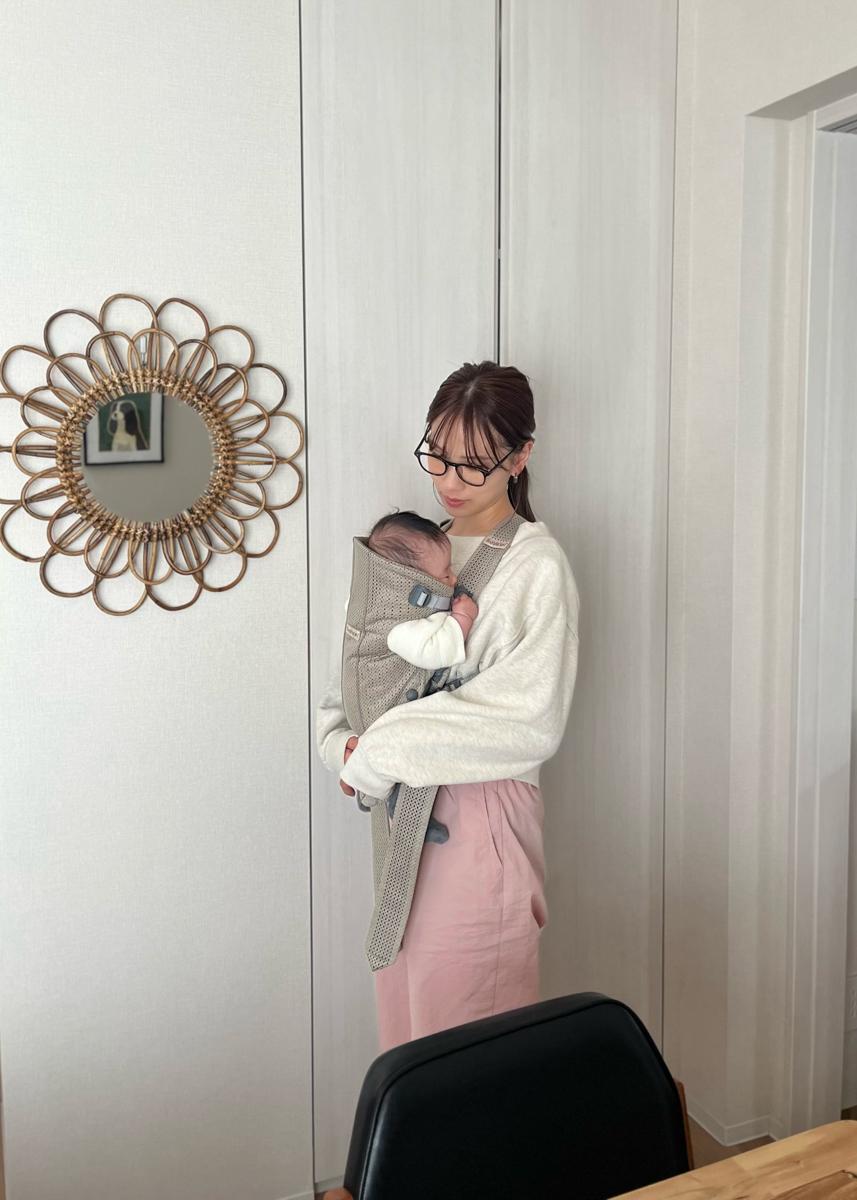 A woman in glasses holds a baby in a carrier, standing by a white wall with a decorative mirror. She wears a white sweater and pink skirt.