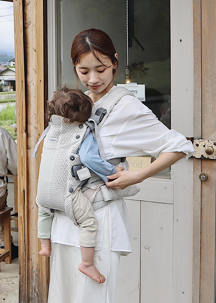 A woman stands outside a building, smiling at a baby in a front carrier. The baby is wearing a blue shirt and light pants.