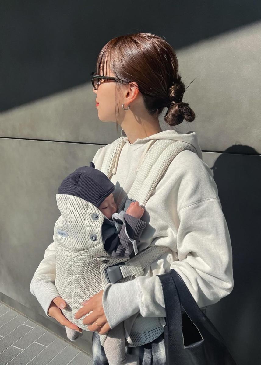 A woman wearing glasses and a hoodie carries a baby in a front carrier. The baby is dressed in a dark outfit and hat.