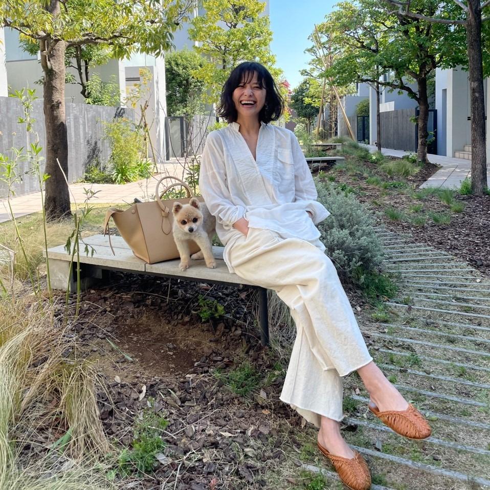 Woman in white outfit sits on a bench with a small dog, surrounded by trees and modern buildings on a sunny day.