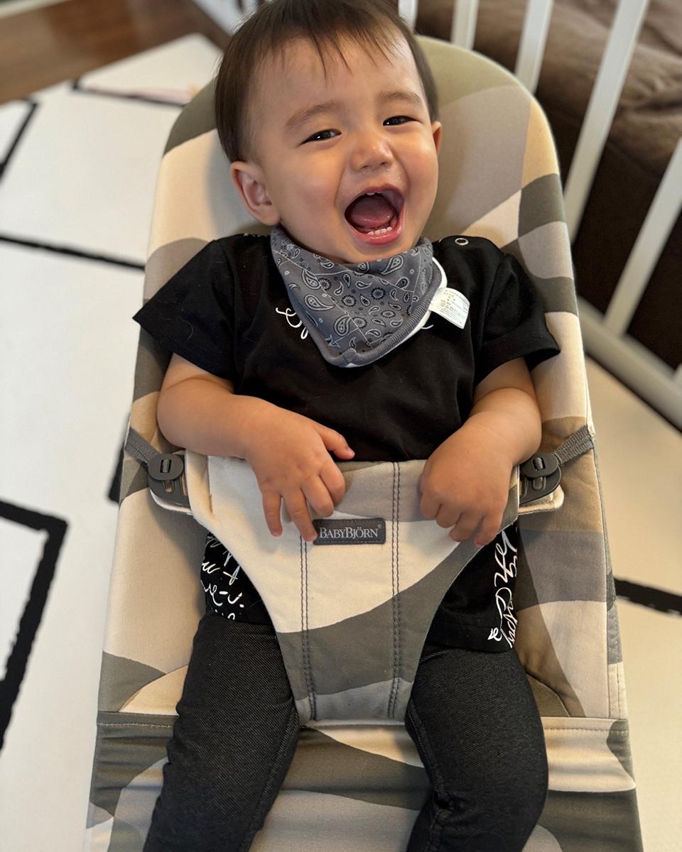 A joyful baby wearing a black outfit and patterned bib sits in a BabyBjörn bouncer, smiling widely.