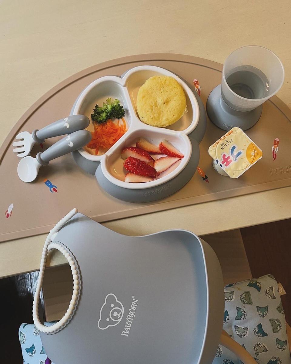 Baby meal setup with divided plate of pancake, strawberries, carrots, broccoli. Includes utensils, water, yogurt, and a bib on a placemat.