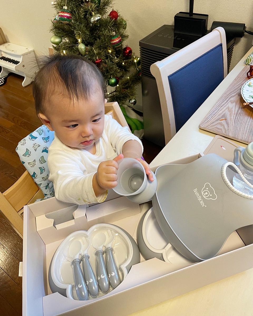 A baby sits at a table, examining a gray cup from a baby dining set. A small Christmas tree is in the background.