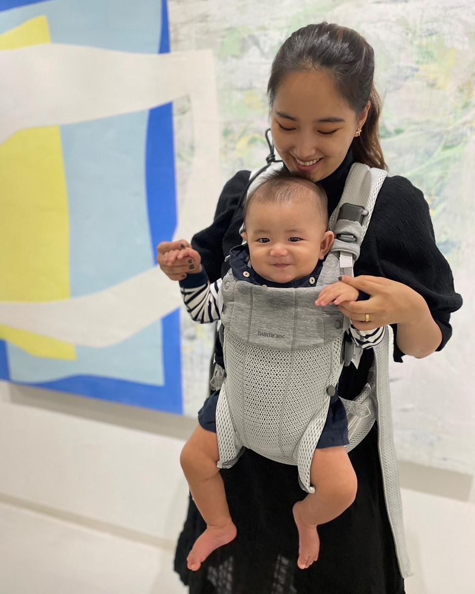 A woman smiles at a baby in a front carrier. The baby is also smiling. They are in front of a colorful abstract painting.