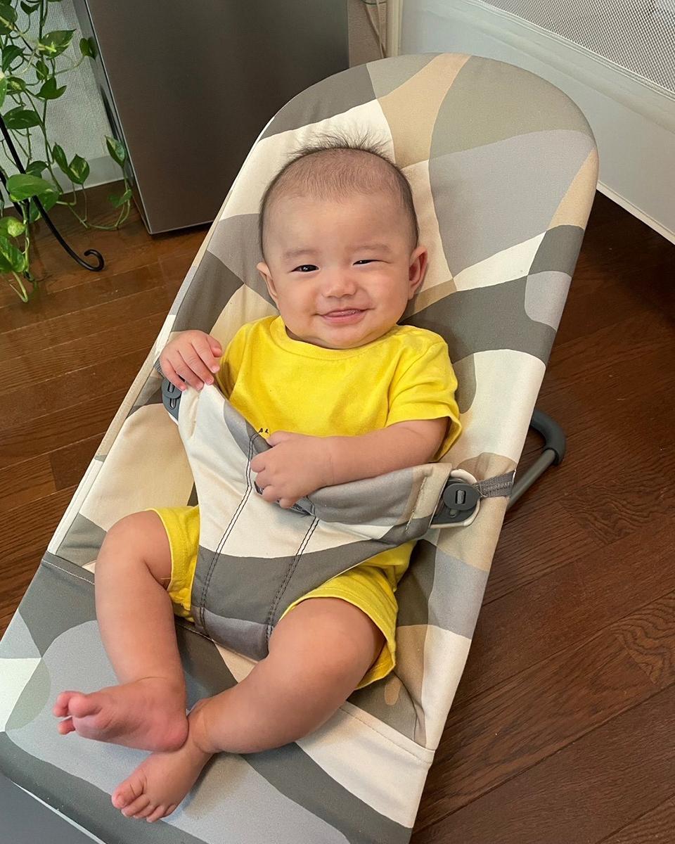 Smiling baby in a yellow outfit sits in a patterned baby bouncer on a wooden floor, with a plant in the background.