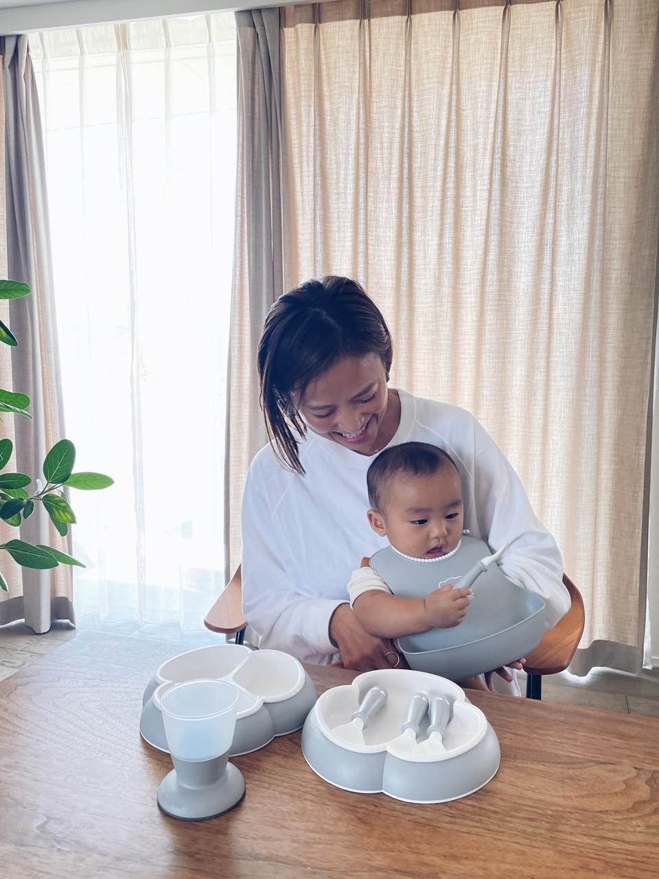 A woman holding a baby in her lap at a dining table with baby dishes and a cup. Sunlight filters through sheer curtains in the background.