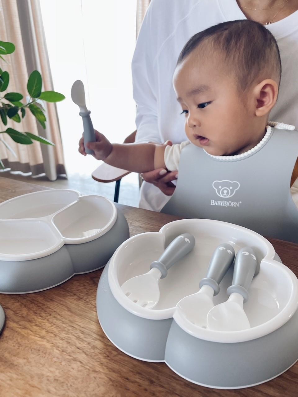 Baby in a bib sits at a table with a divided plate and utensils, holding a spoon. An adult helps guide the child's hand.