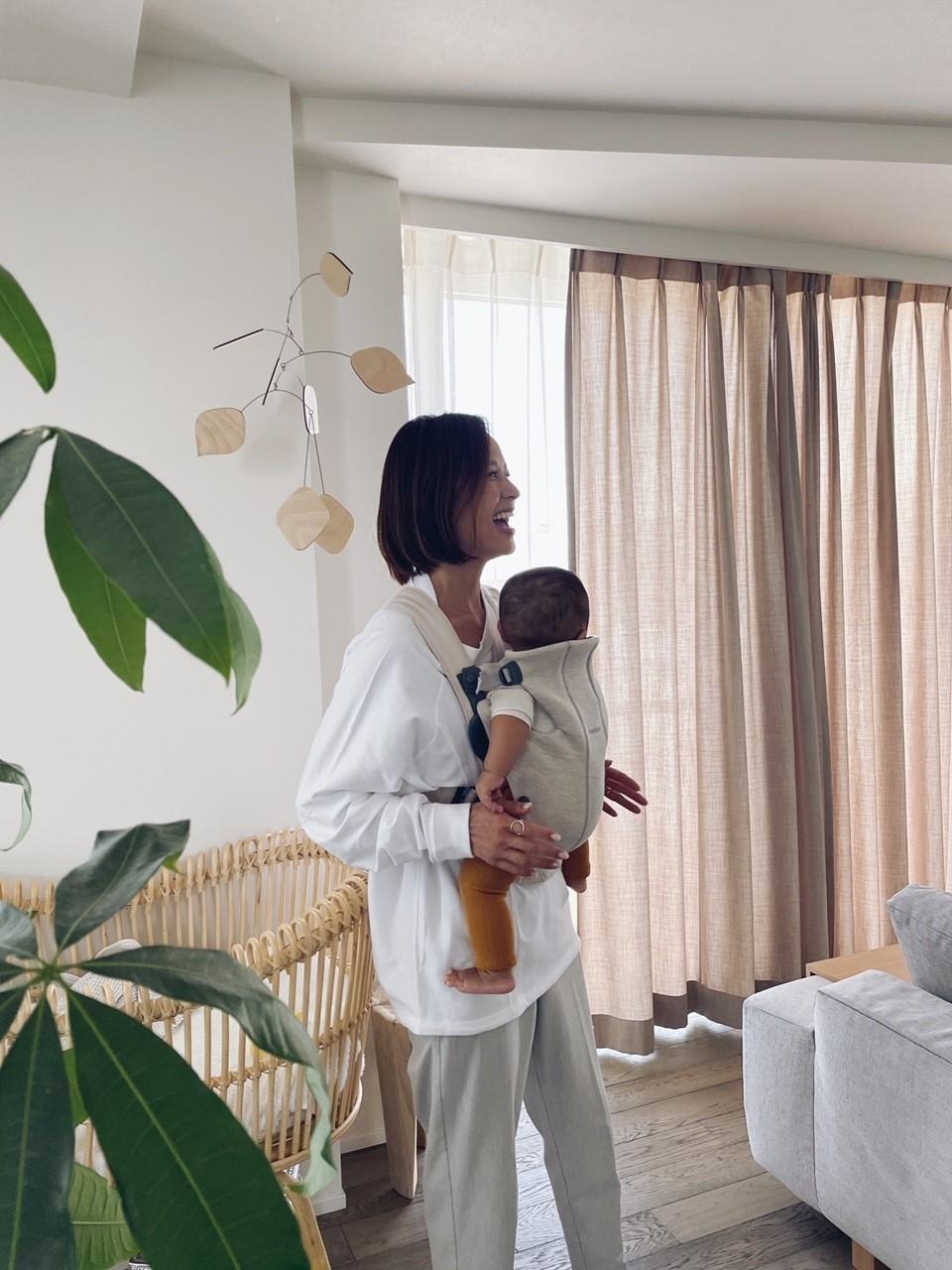 A woman stands in a bright room, smiling while holding a baby in a carrier. Nearby are a crib, a large plant, and sheer curtains.