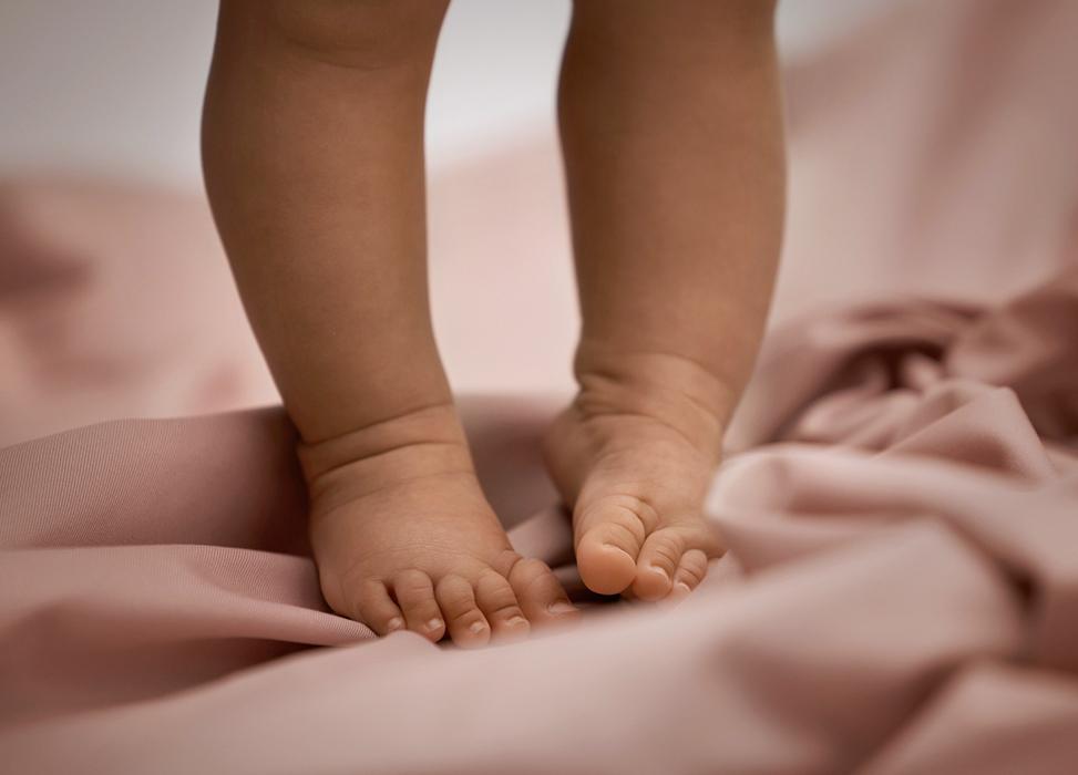 Close-up of a baby's feet standing on soft, pink fabric, with focus on tiny toes and smooth skin.