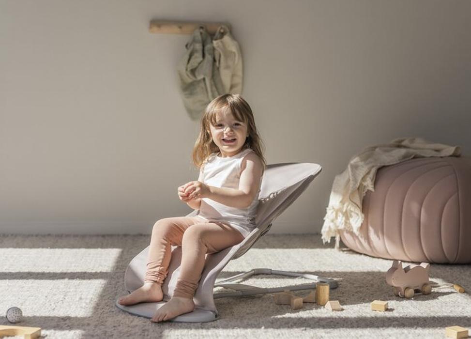 A smiling child sits in a soft chair on a sunlit carpeted floor, surrounded by wooden toys, with a pouf and hanging clothes in the background.
