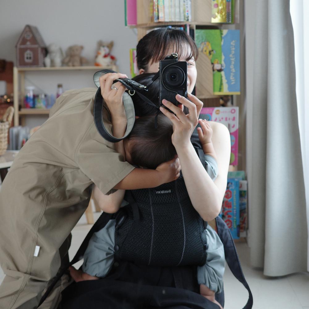 A person takes a mirror selfie holding a camera, with a child hugging them. Books and toys are visible in the background.
