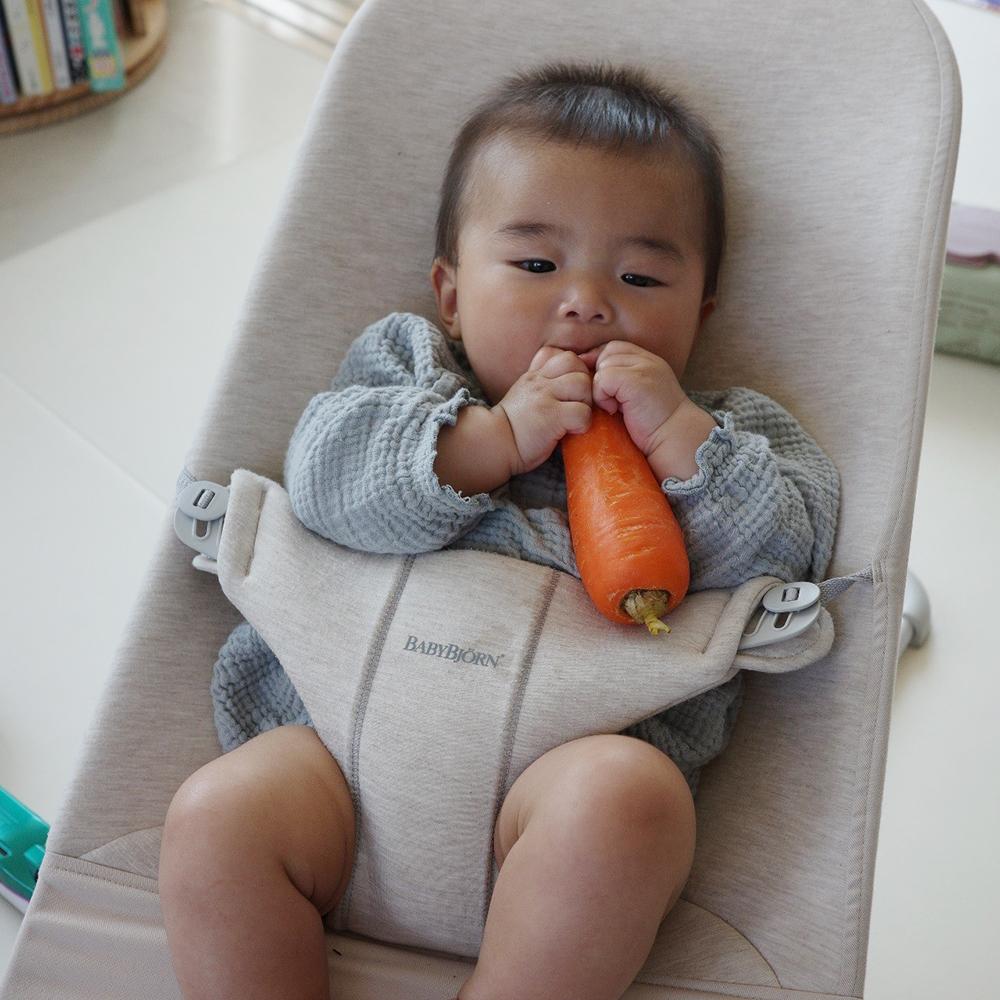 Baby in a bouncer seat holding and chewing on a large carrot, wearing a textured gray outfit.
