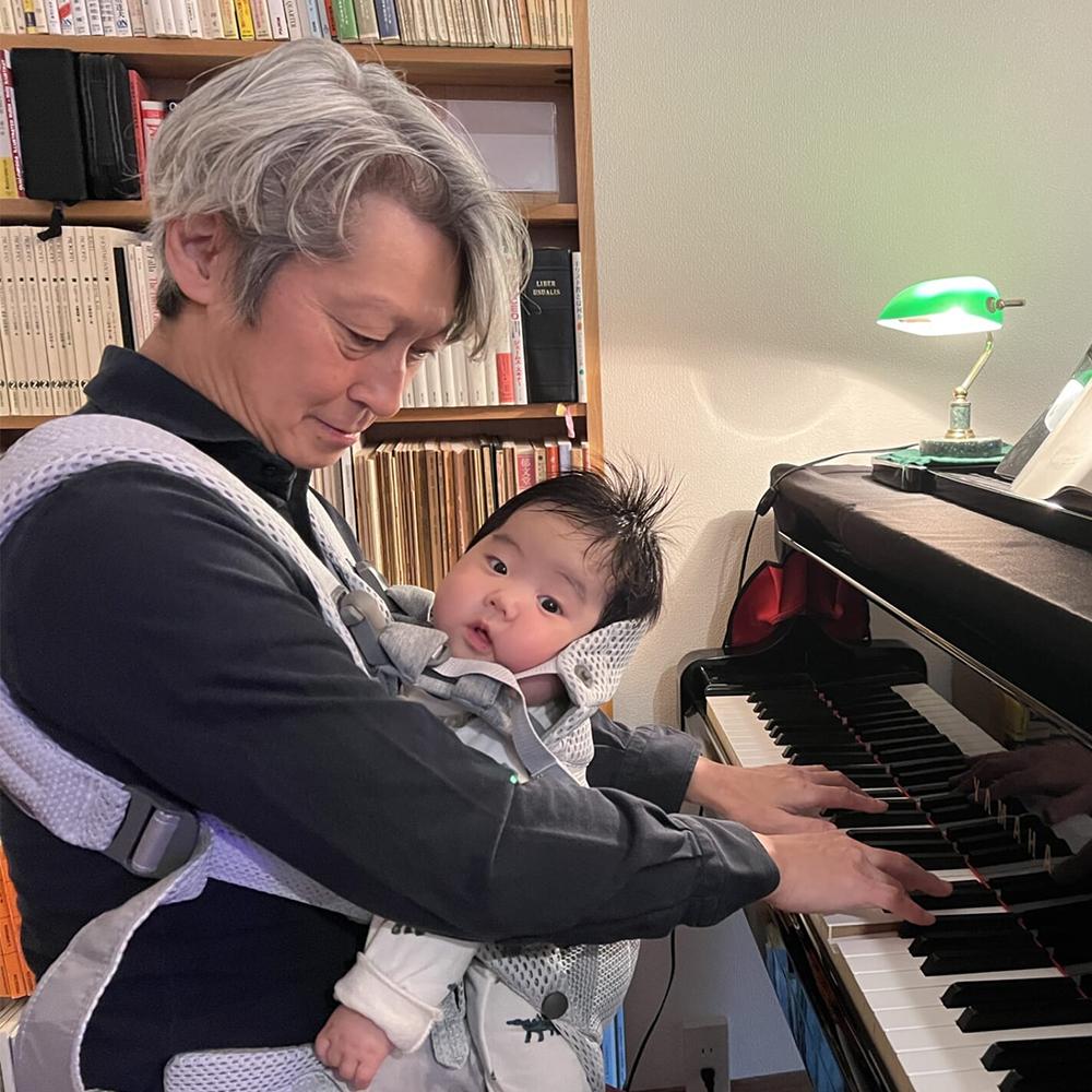 An adult with gray hair plays the piano while carrying a baby in a front carrier. The baby looks at the camera. Books and a lamp are in the background.