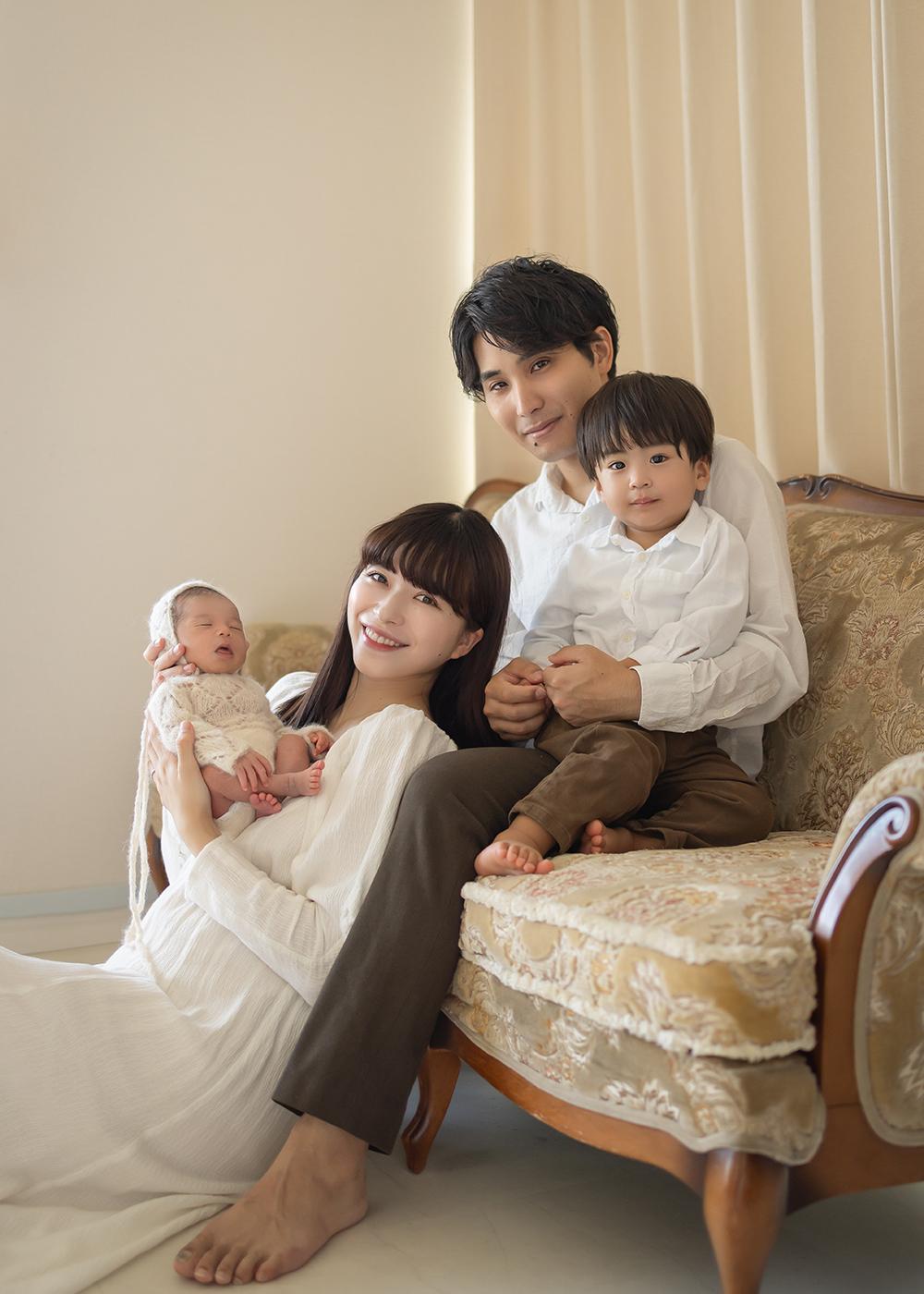 A family of four sits on a vintage sofa. The mother holds a newborn, while the father holds a toddler. All are smiling and dressed in white.