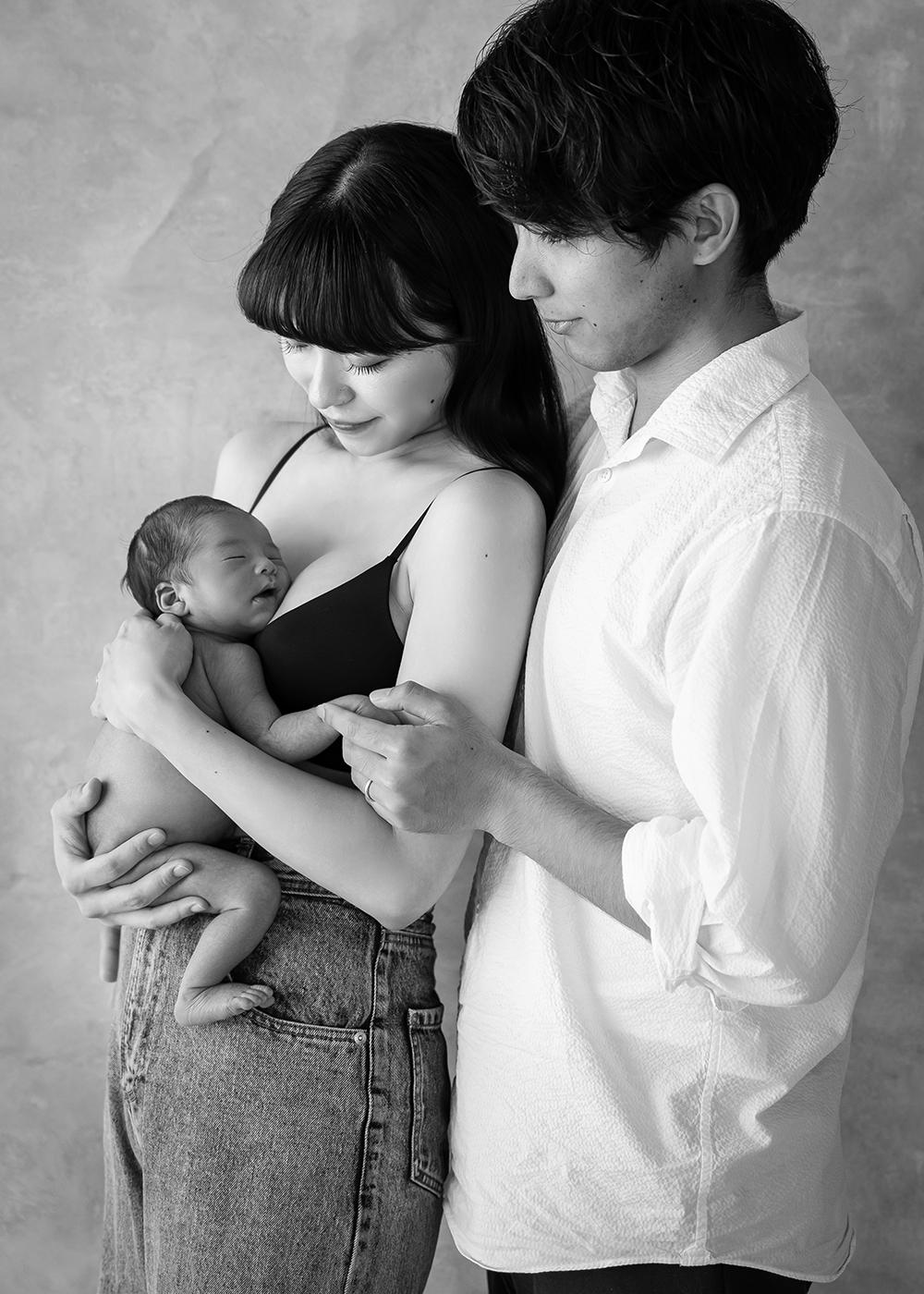 A couple lovingly gazes at their newborn baby, cradled in the mother's arms. The father gently holds the mother's hand. Black and white photo.