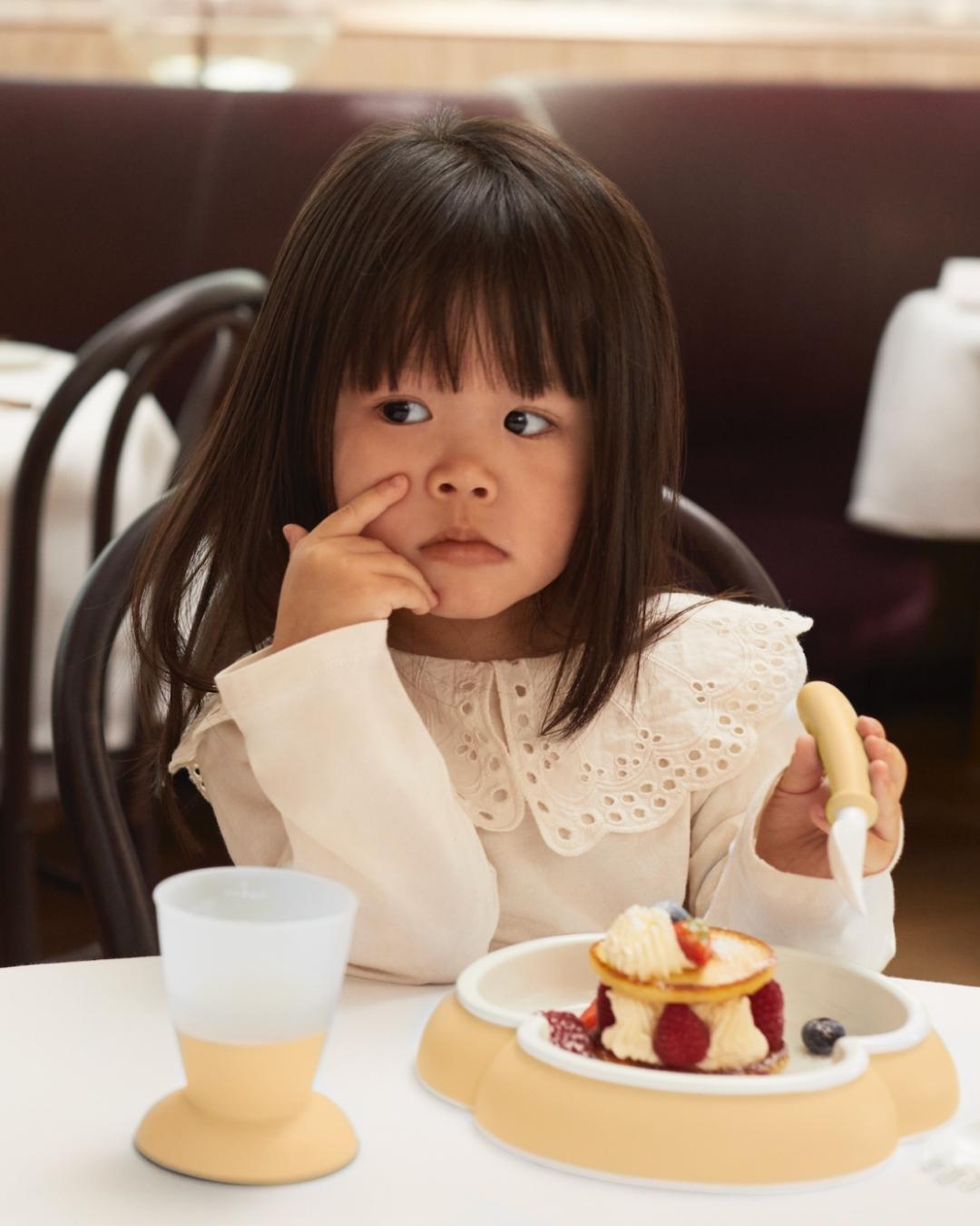A young girl in a white dress sits at a table with a small dessert and cup, looking thoughtful, and holding a fork.