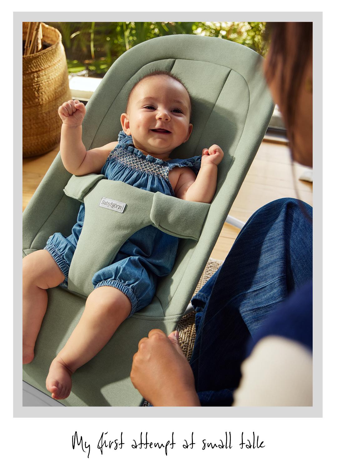 Smiling baby in a green bouncer chair, wearing a blue romper, with an adult nearby. Handwritten text: "My first attempt at small talk."