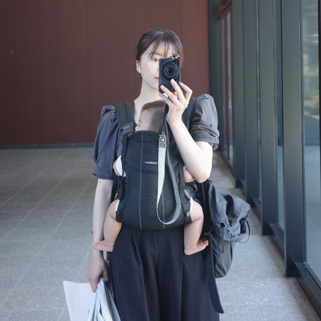 Woman taking a mirror selfie with a camera, carrying a baby in a front-facing carrier, standing in a modern indoor setting.