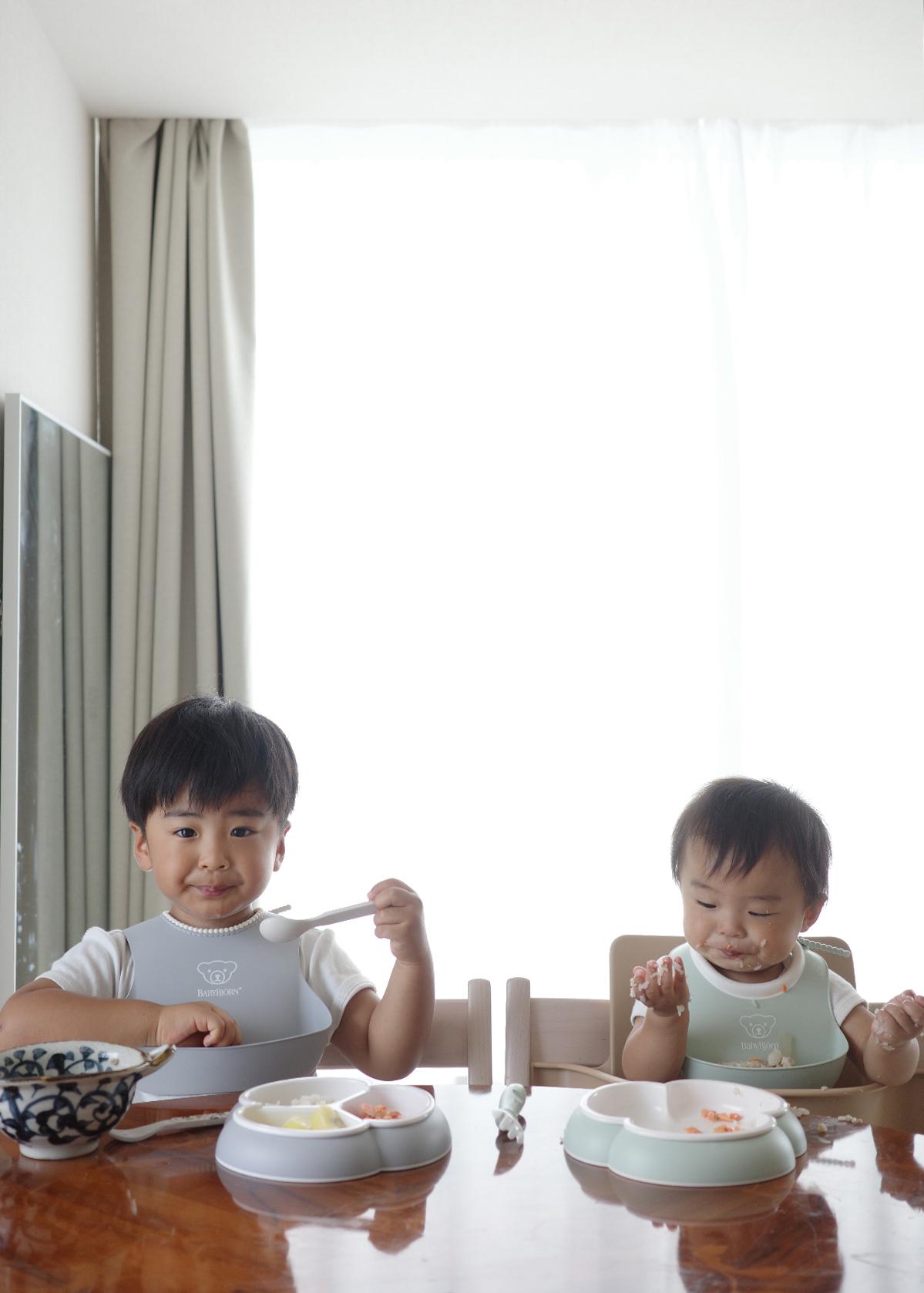 Two young children wearing bibs sit at a table eating from bowls, with food on their faces, in a bright room with a large window.
