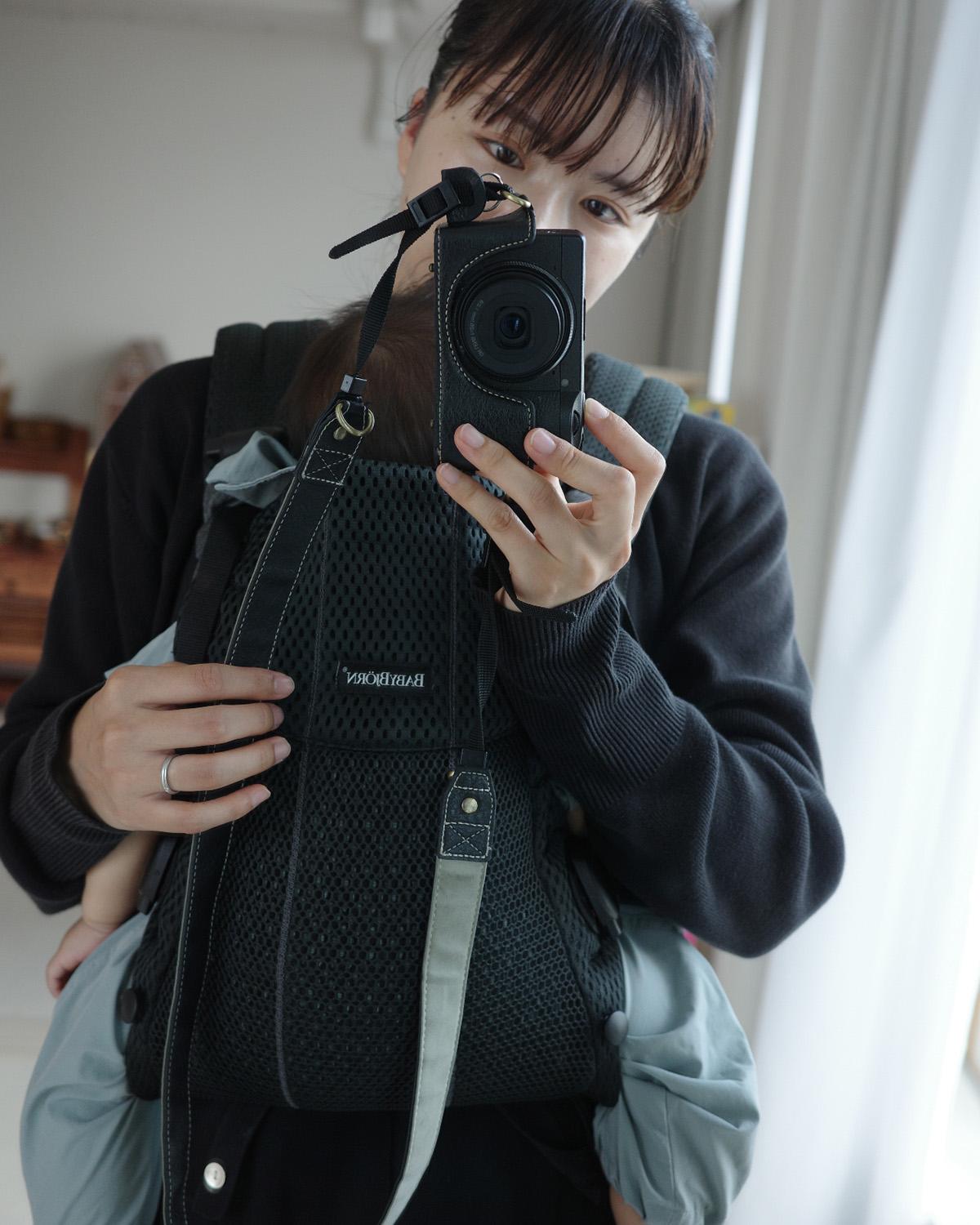 Person taking a mirror selfie while holding a baby in a black carrier, wearing a dark sweater, in a softly lit room.