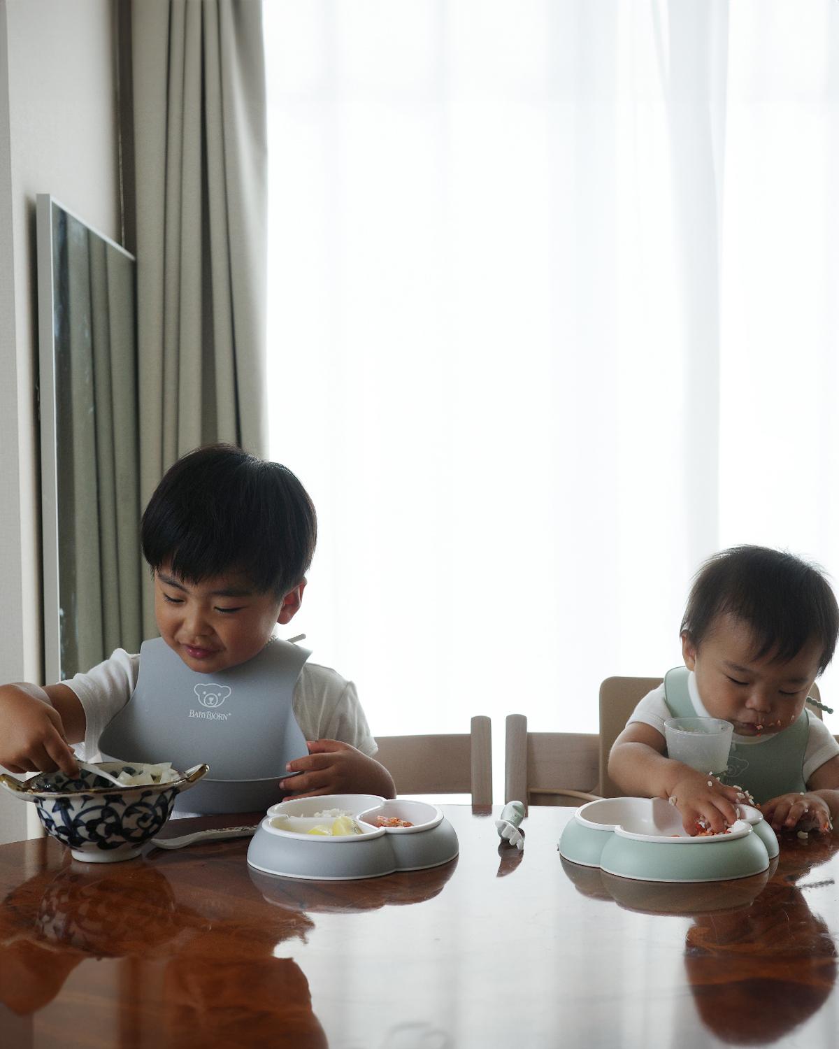 Two young children sitting at a table, wearing bibs, eating from divided plates. The older child uses a spoon, while the younger uses hands.