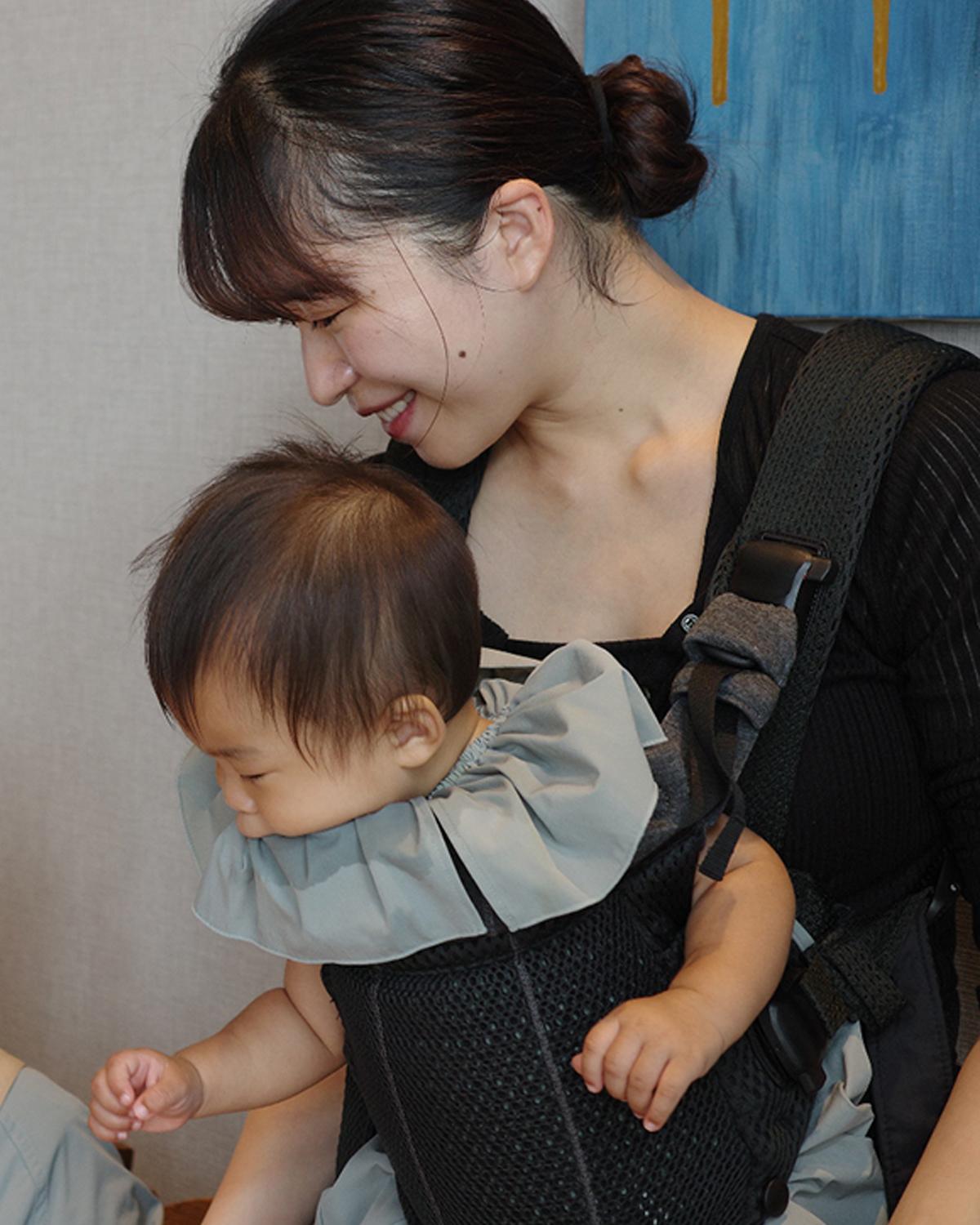 A woman smiles while holding a baby in a front carrier. Both are wearing matching outfits. The background features a blue and beige wall.