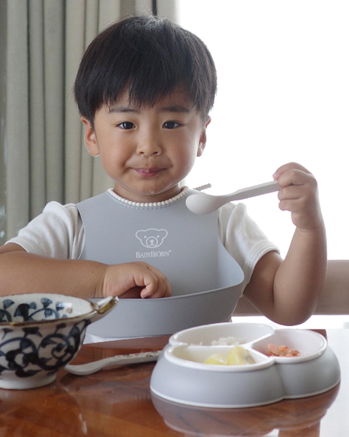 A young child with a bib is sitting at a table, holding a spoon and eating from a divided plate with various foods.
