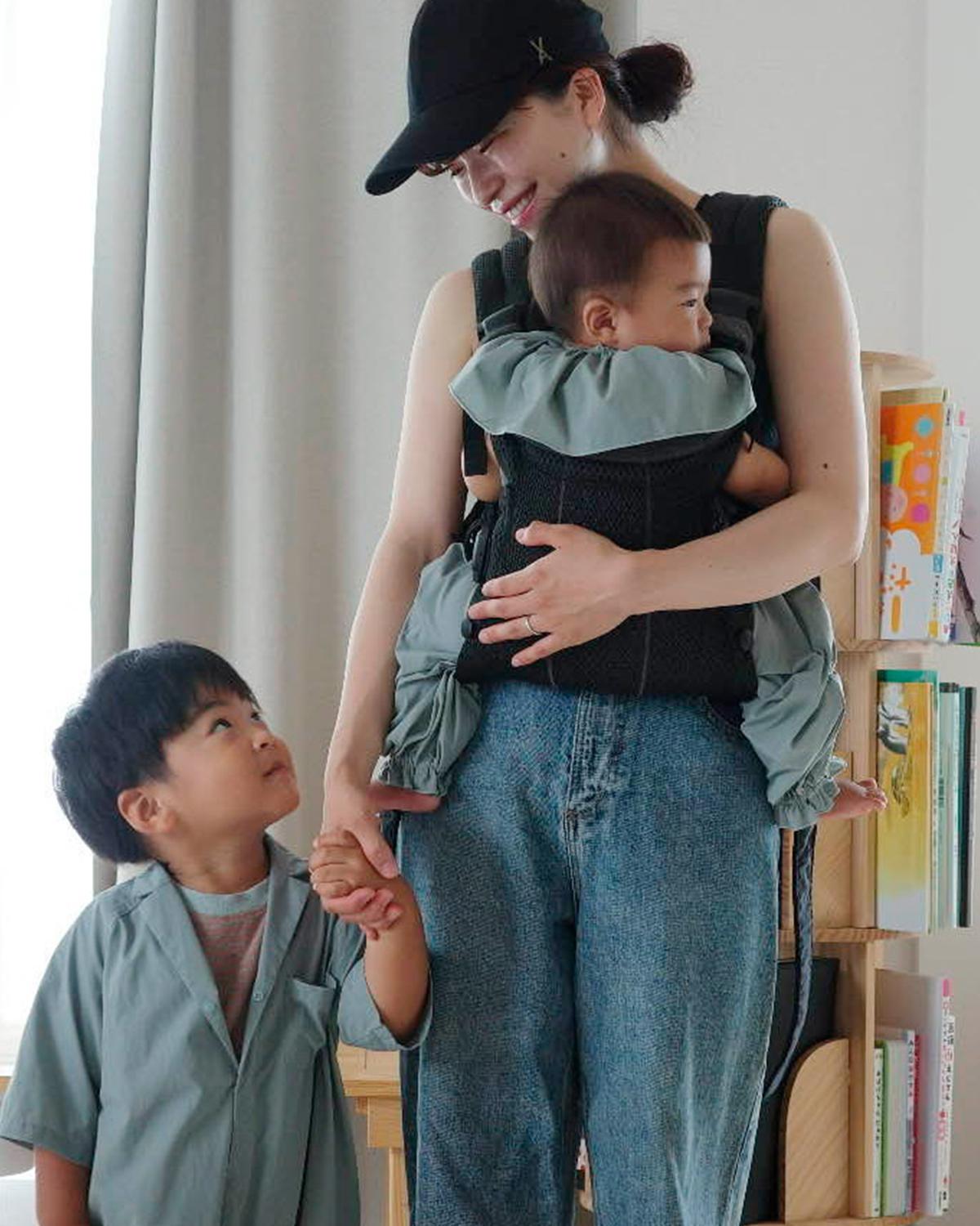 A woman wearing a cap holds a baby in a carrier while holding hands with a young child. Both children wear matching outfits.
