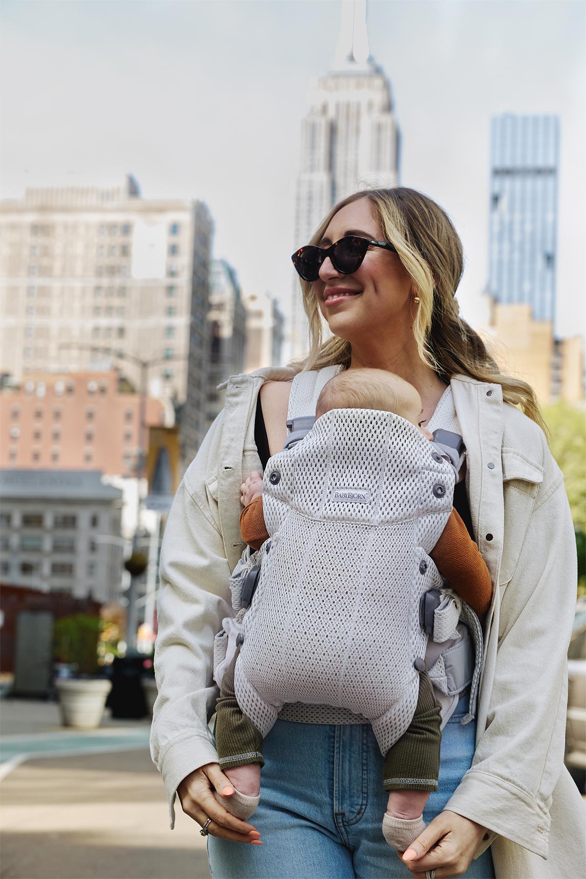 A woman wearing sunglasses carries a baby in a gray carrier. They are outdoors in a city with tall buildings in the background.