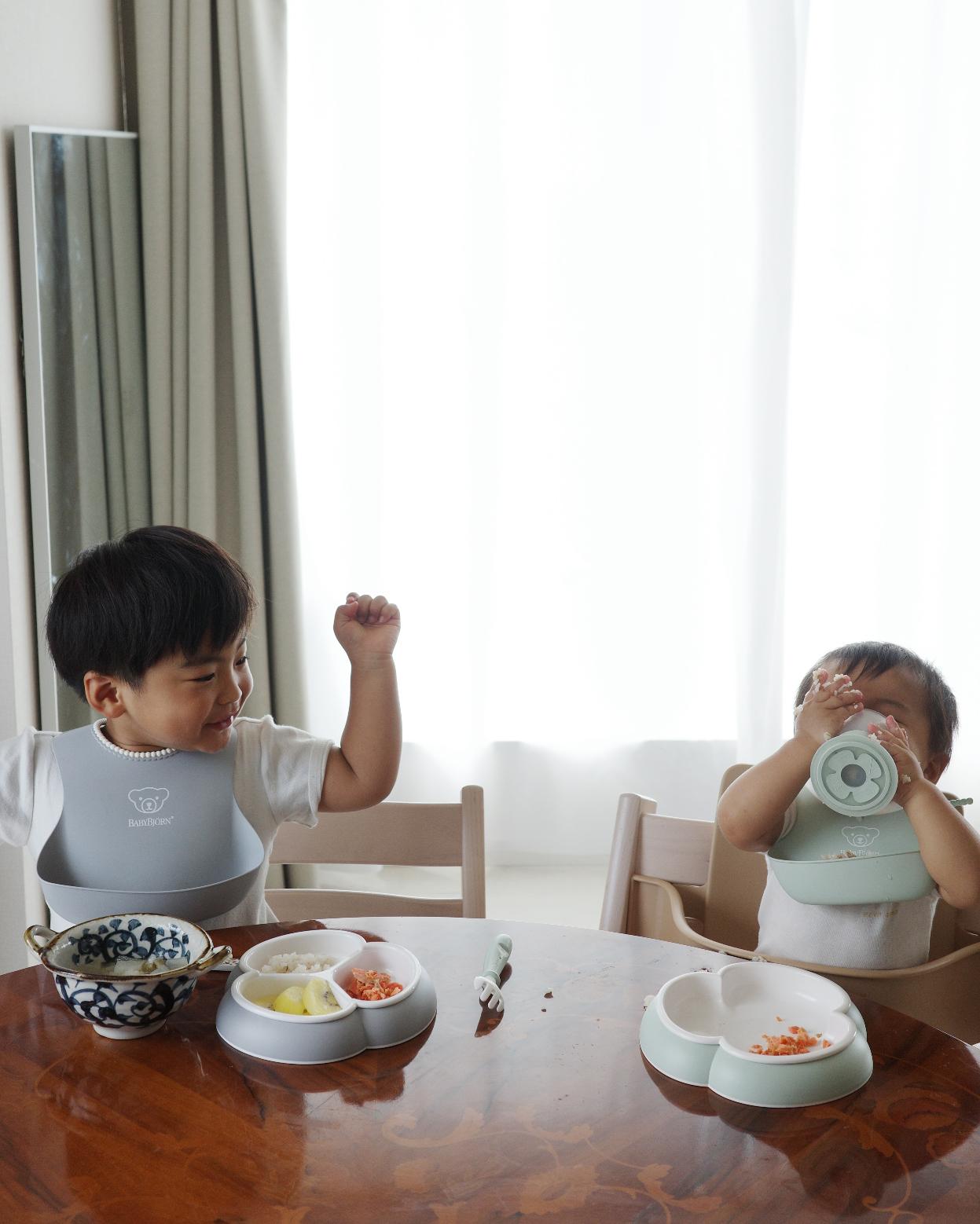 Two young children sitting at a table, wearing bibs, enjoying a meal with divided plates and bowls, in a bright room.