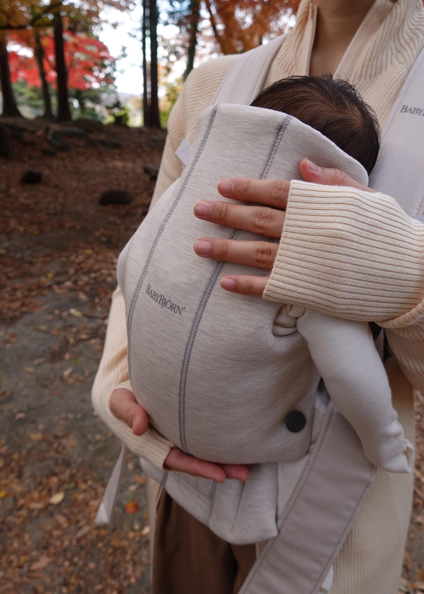 Person wearing a beige sweater holds a baby in a light gray carrier outdoors, surrounded by autumn leaves.