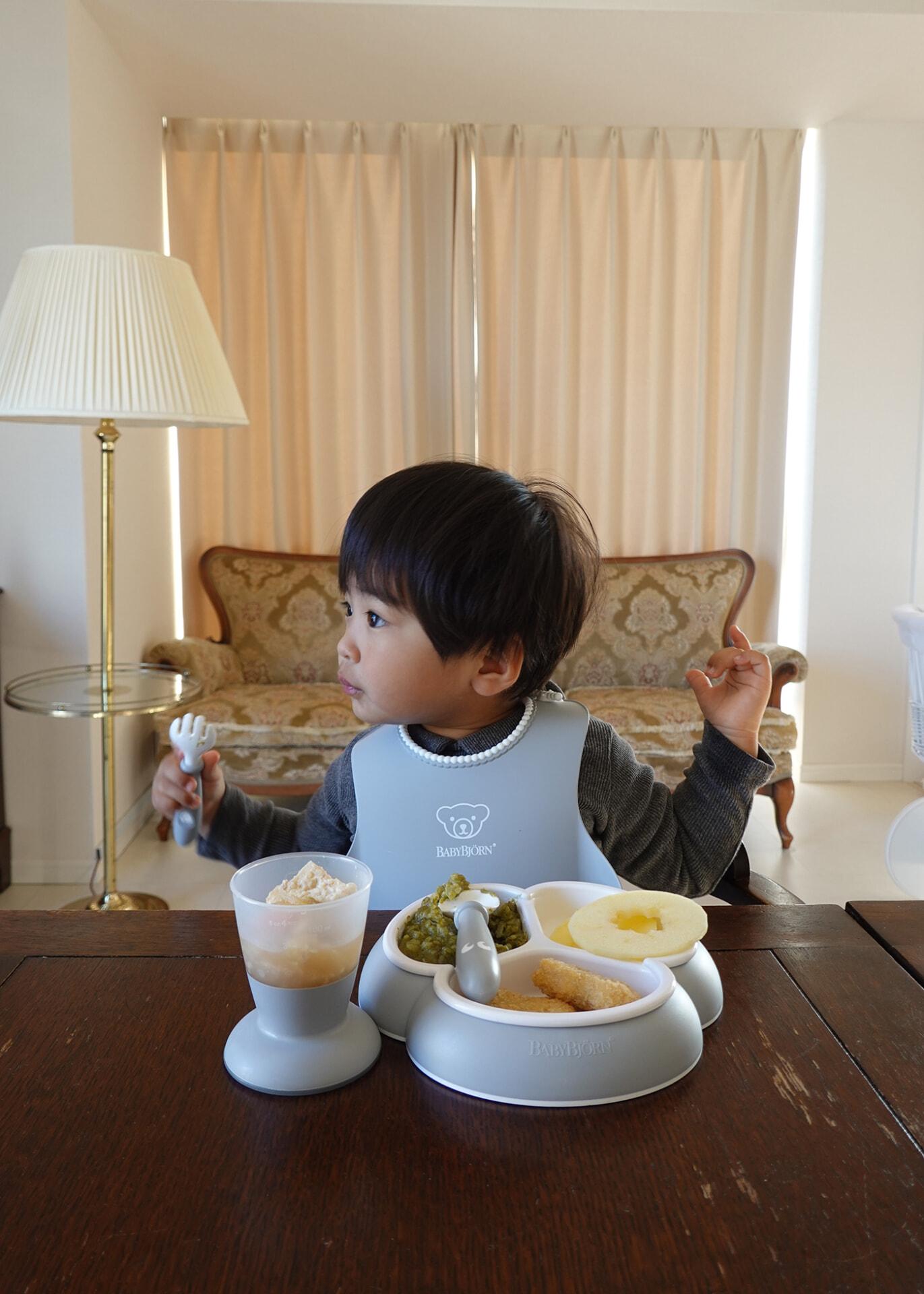 A toddler sits at a table with a divided plate of food, wearing a bib. The room has a sofa, lamp, and curtains in the background.