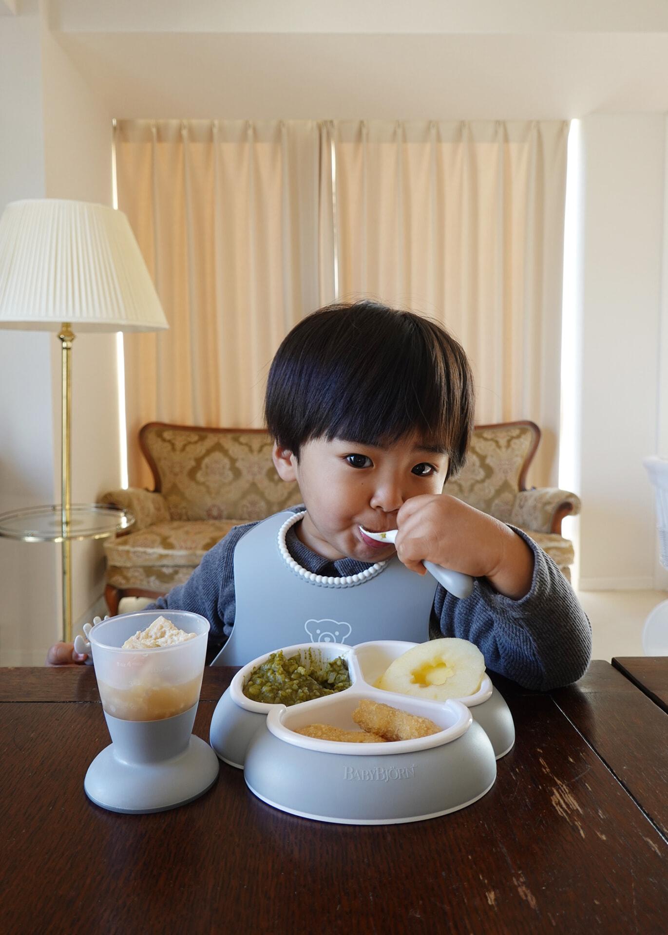 A young child eating with a spoon, wearing a bib, sits at a table with cups of food in front, in a warmly lit room with curtains.