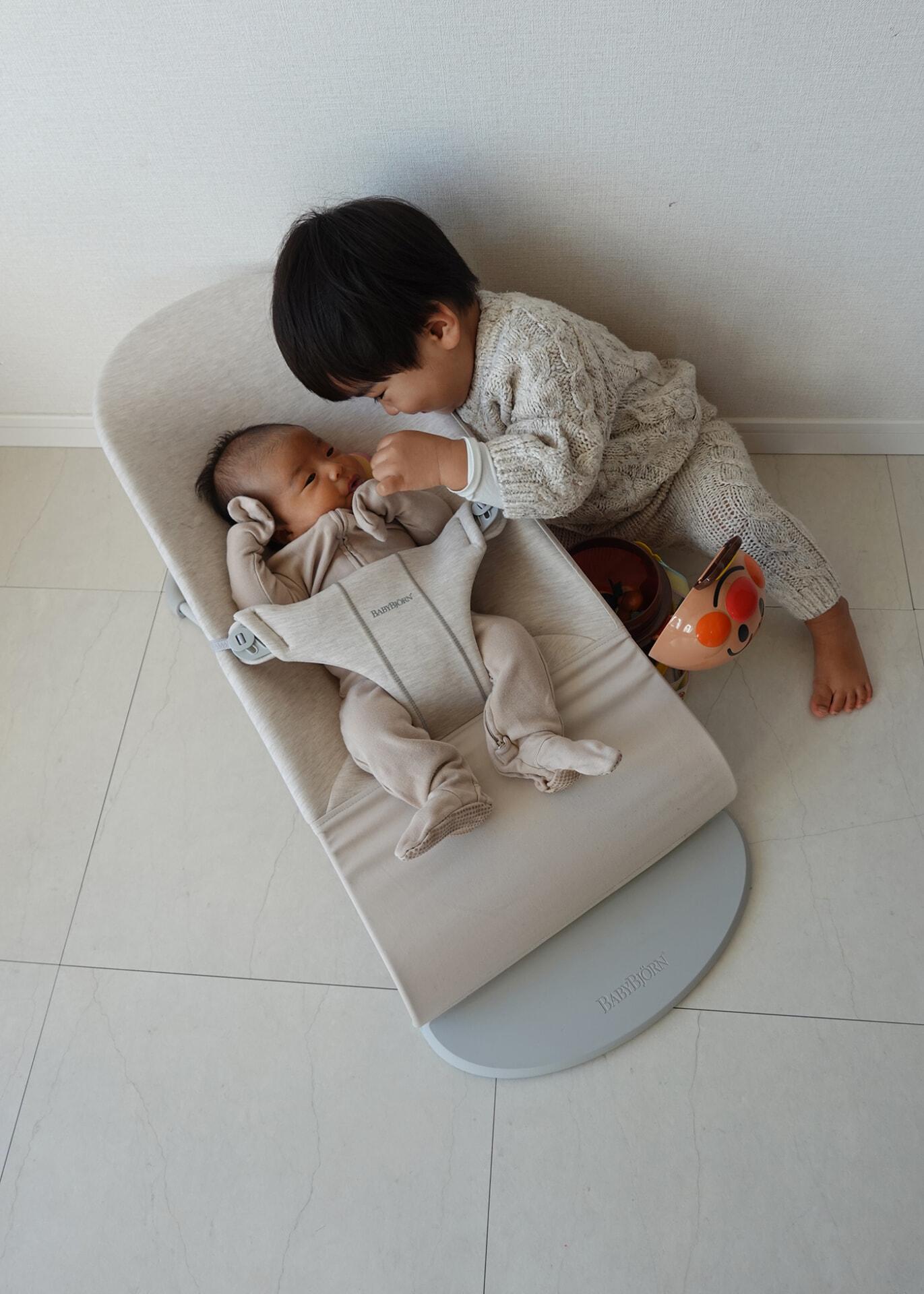 A toddler gently touches a baby lying in a beige baby bouncer. Both are dressed in cozy outfits on a light-colored floor.