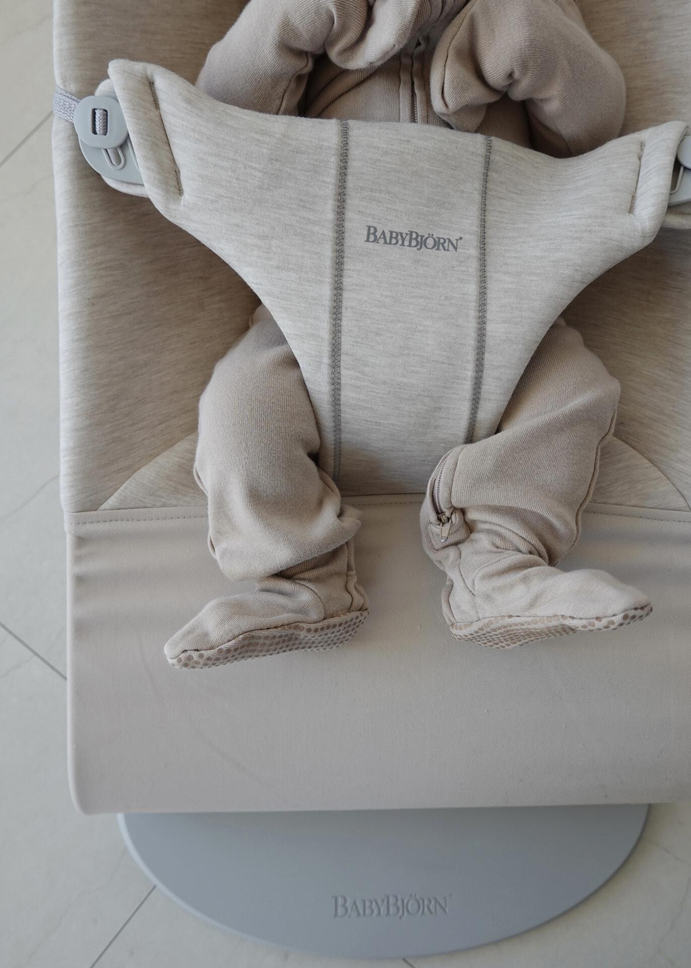 Baby in a beige BabyBjörn bouncer, wearing a matching outfit with footed pants, partially visible from the neck down.