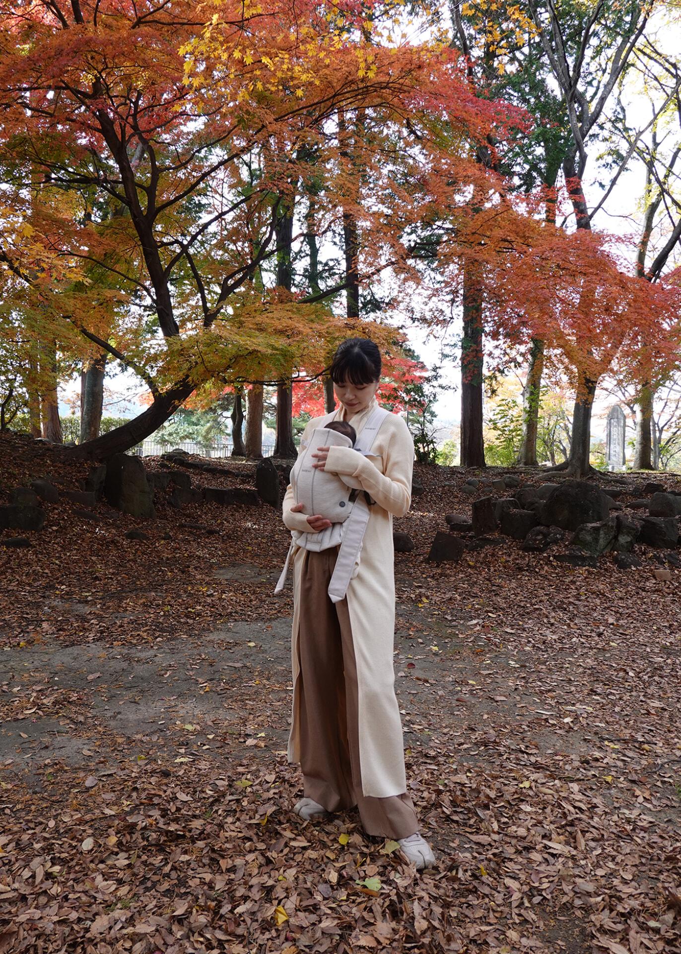 A person stands on a leaf-covered path, holding a baby, surrounded by vibrant autumn trees with red and orange leaves.