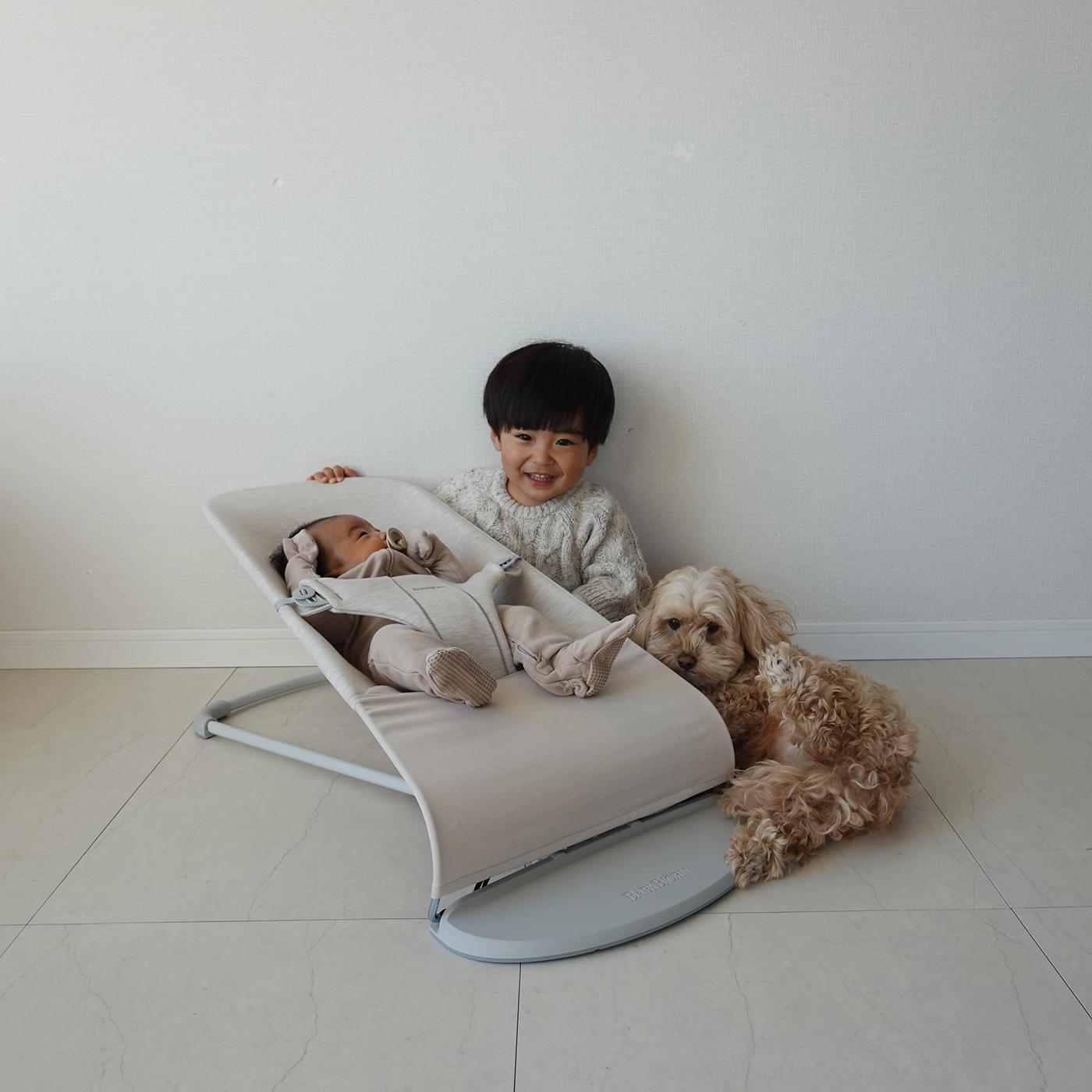 A child smiles next to a baby in a bouncer and a fluffy dog lying on the floor, all against a plain white wall.