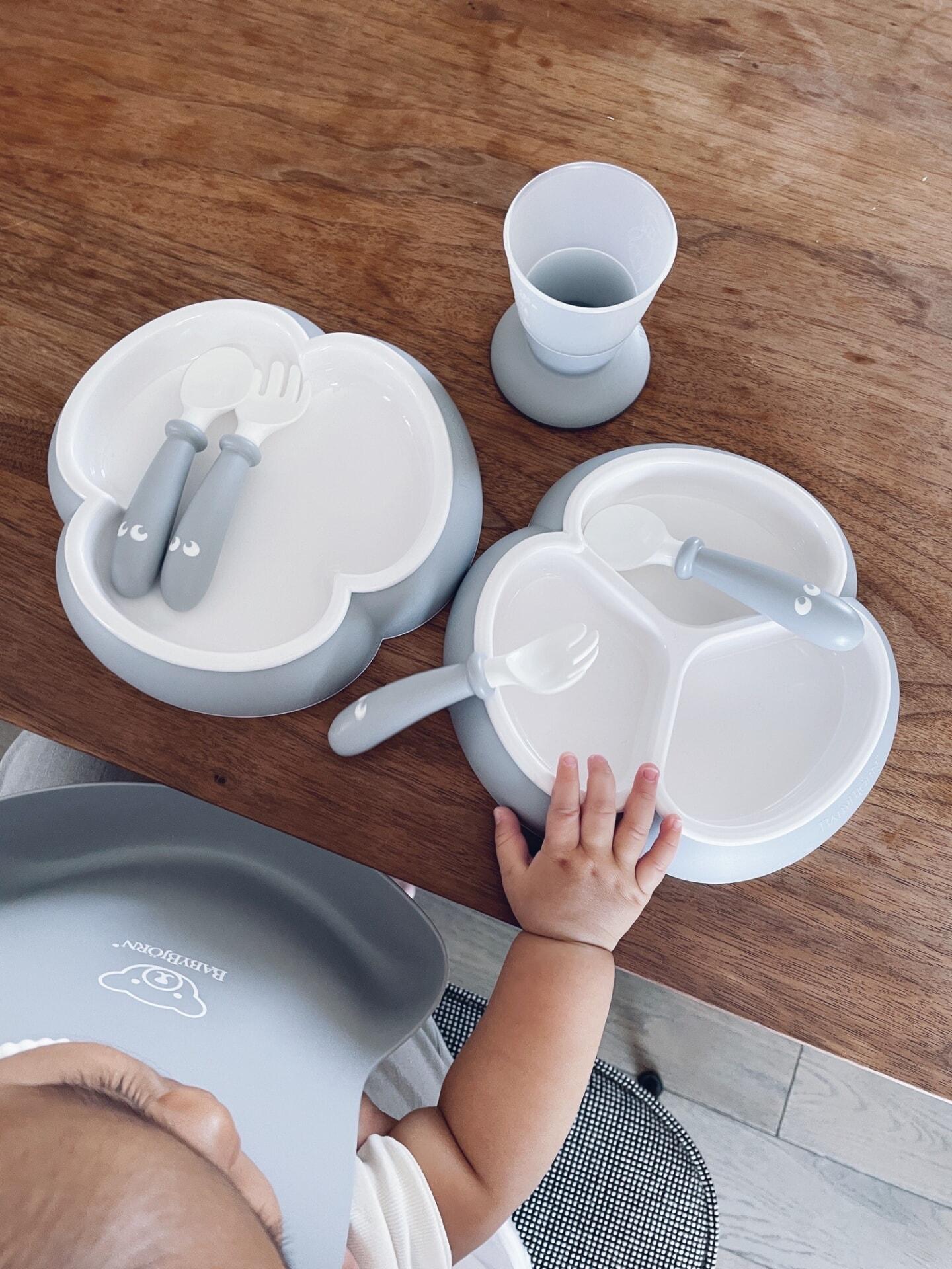 A baby reaches for a set of white and gray divided plates with matching utensils on a wooden table, next to a gray cup.
