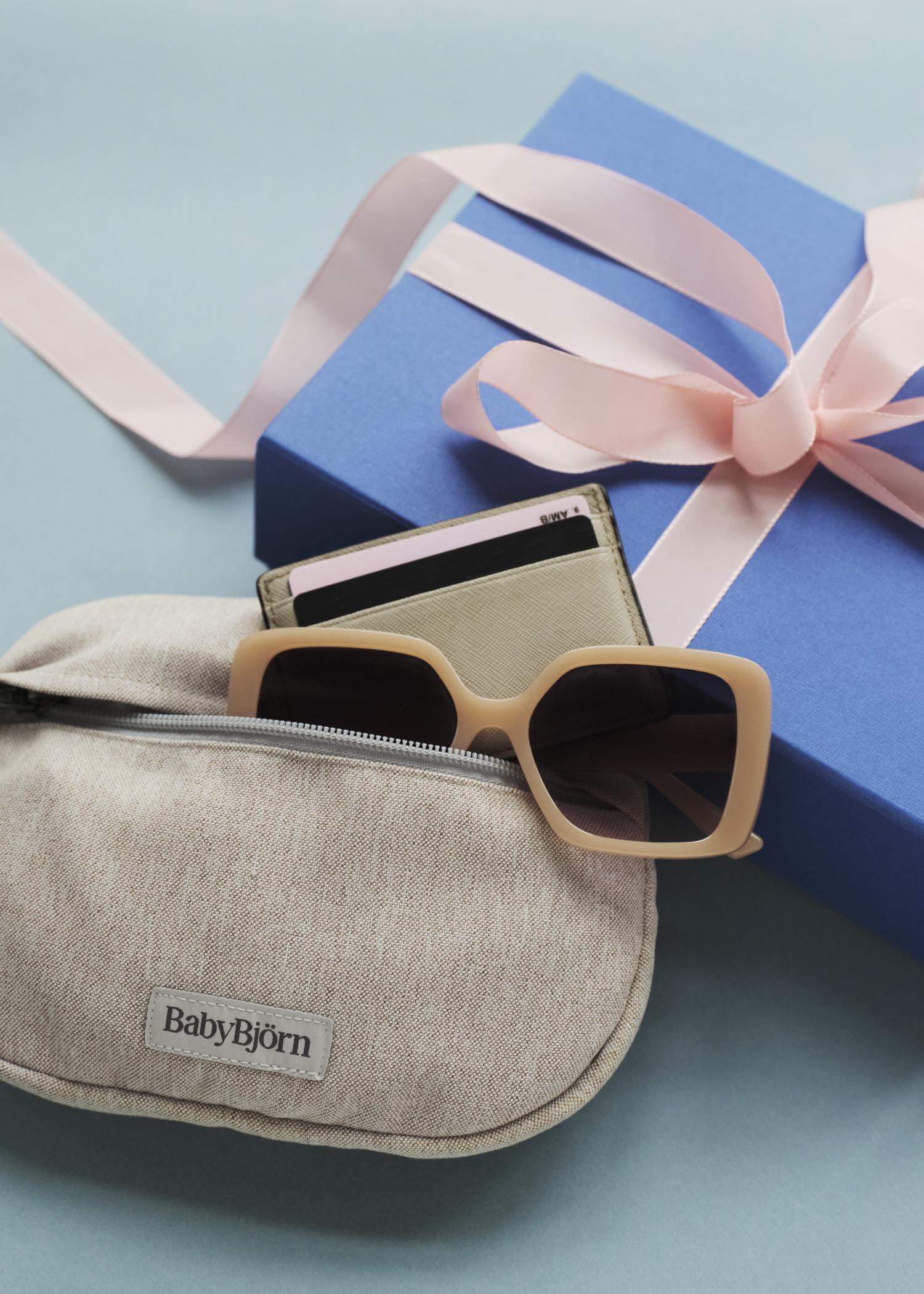 A blue gift box with a pink ribbon next to a beige pouch containing sunglasses and a wallet on a light blue surface.