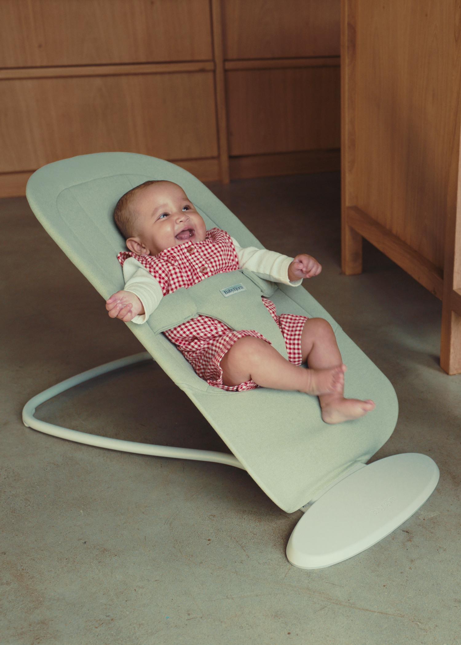 Smiling baby in a red checkered outfit sits in a light green bouncer on the floor, with wooden cabinets in the background.