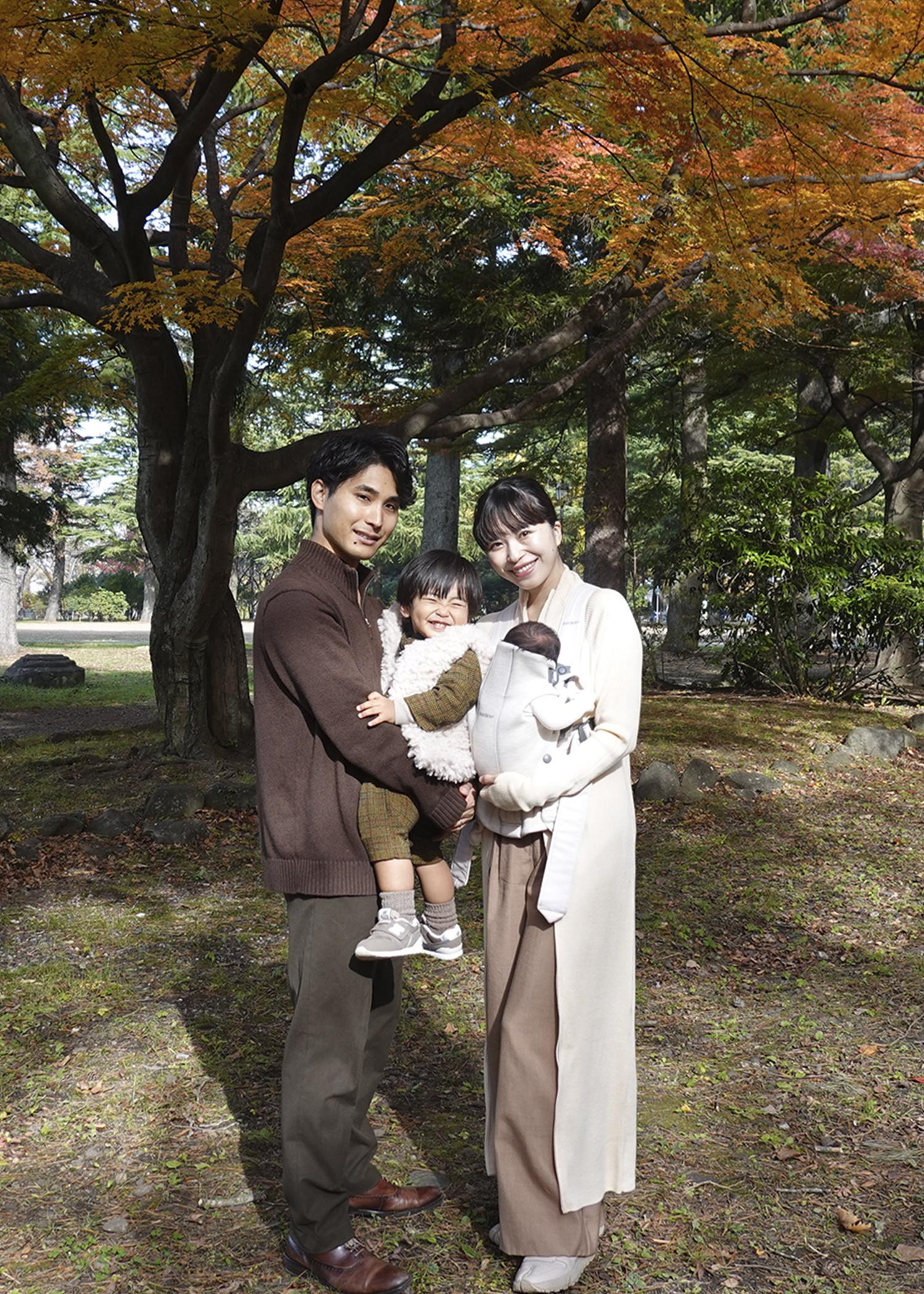 A family of three stands in a park with autumn leaves, smiling and posing for a photo under a tree.