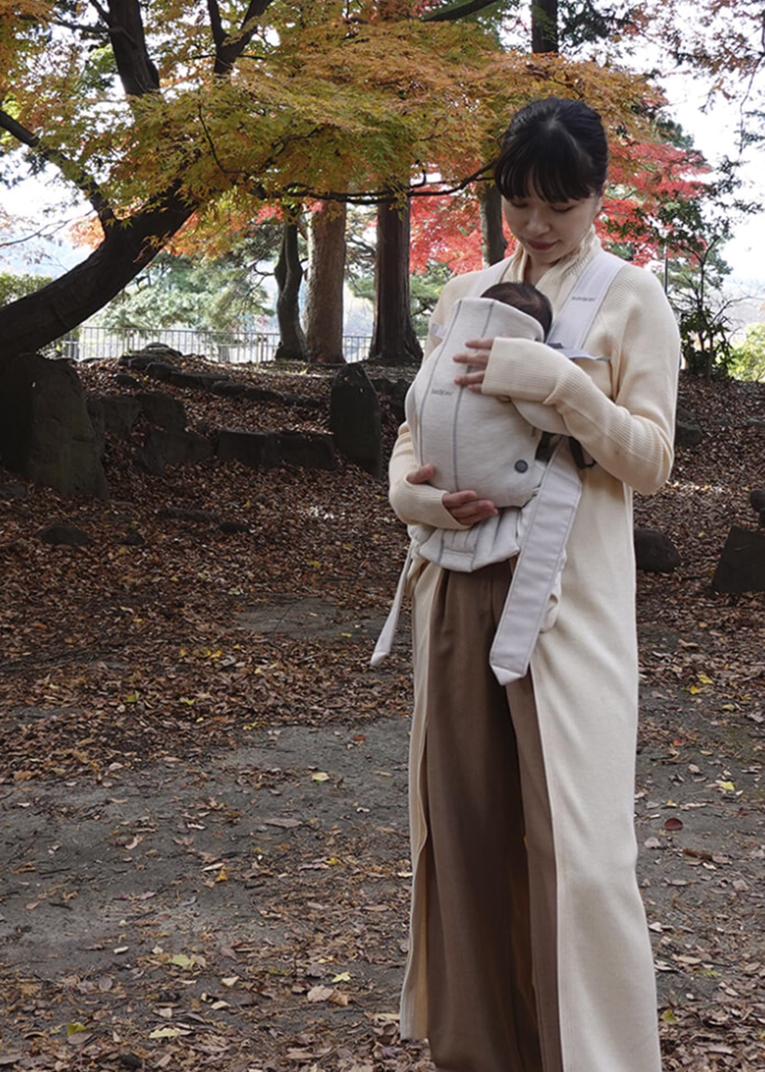 A woman stands in a park with autumn leaves, holding a baby in a carrier. She wears a long coat and looks down at the baby affectionately.