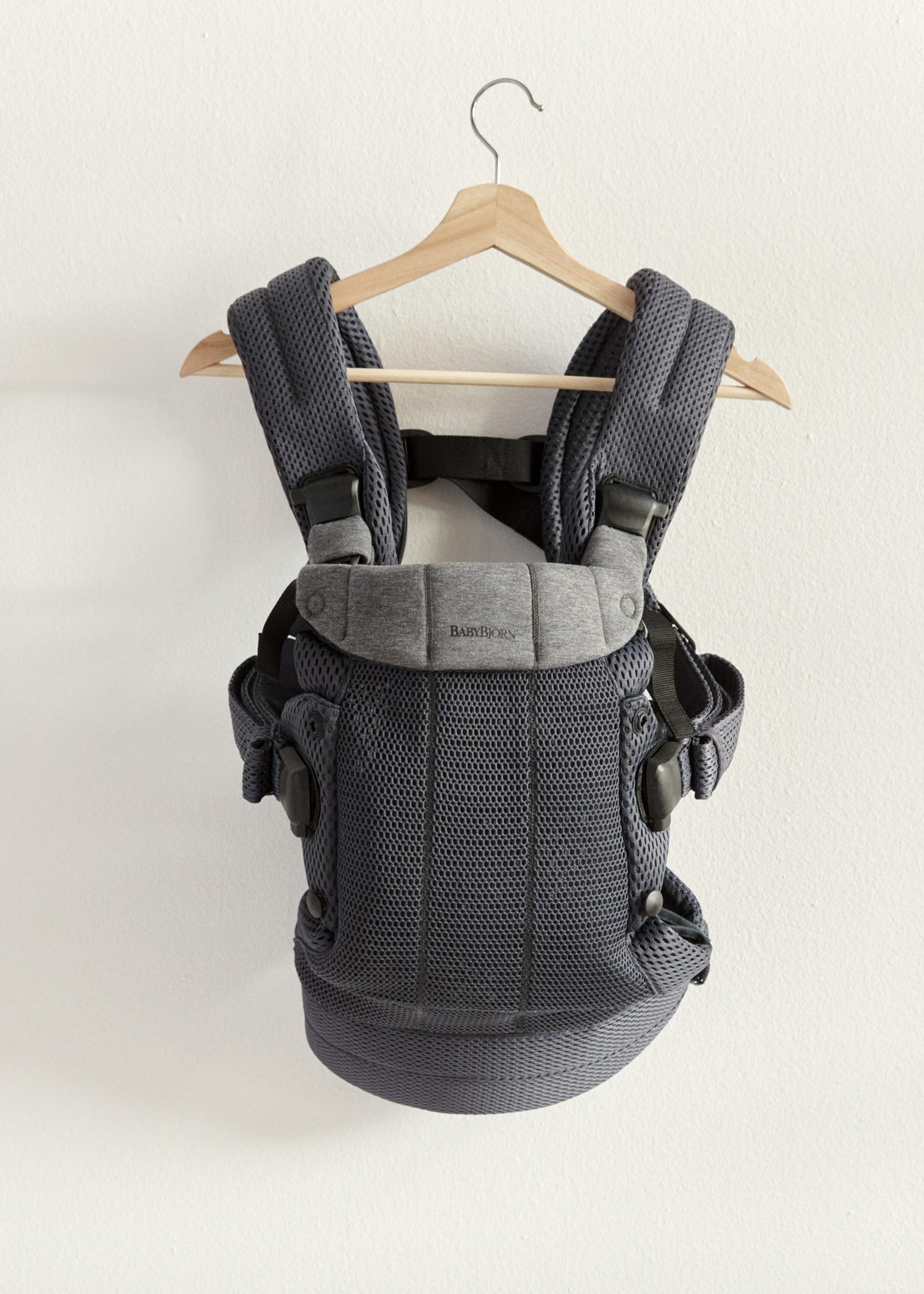 Gray baby carrier with padded straps and adjustable buckles, hanging on a wooden hanger against a white wall.