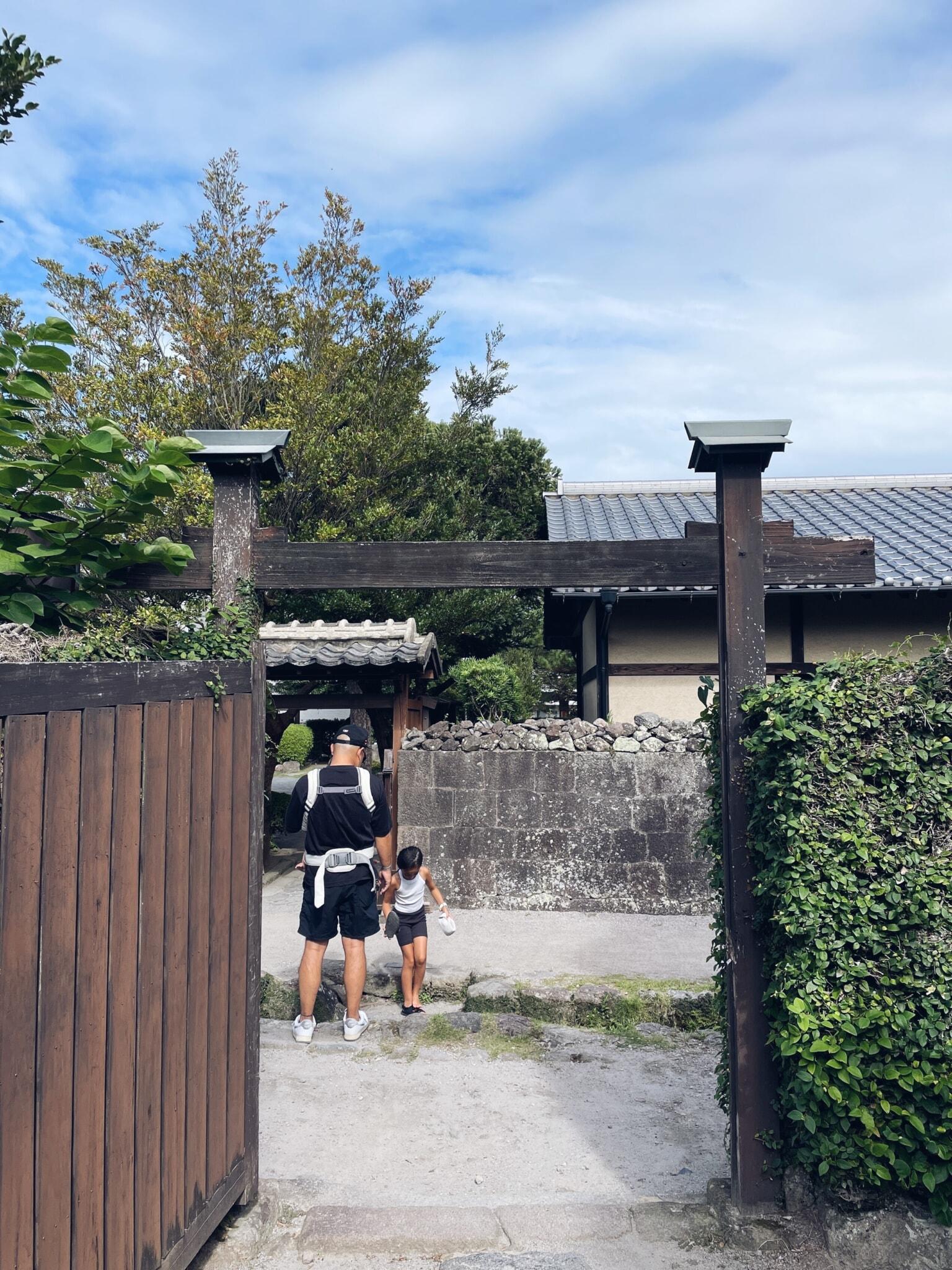 Dad wearing baby carrier and toddler walk through a wooden gate towards a traditional building, surrounded by greenery under a cloudy sky.