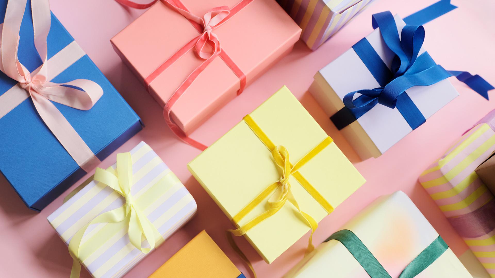 Colorful gift boxes with various ribbons arranged on a pink surface, featuring shades of blue, pink, yellow, and striped patterns.