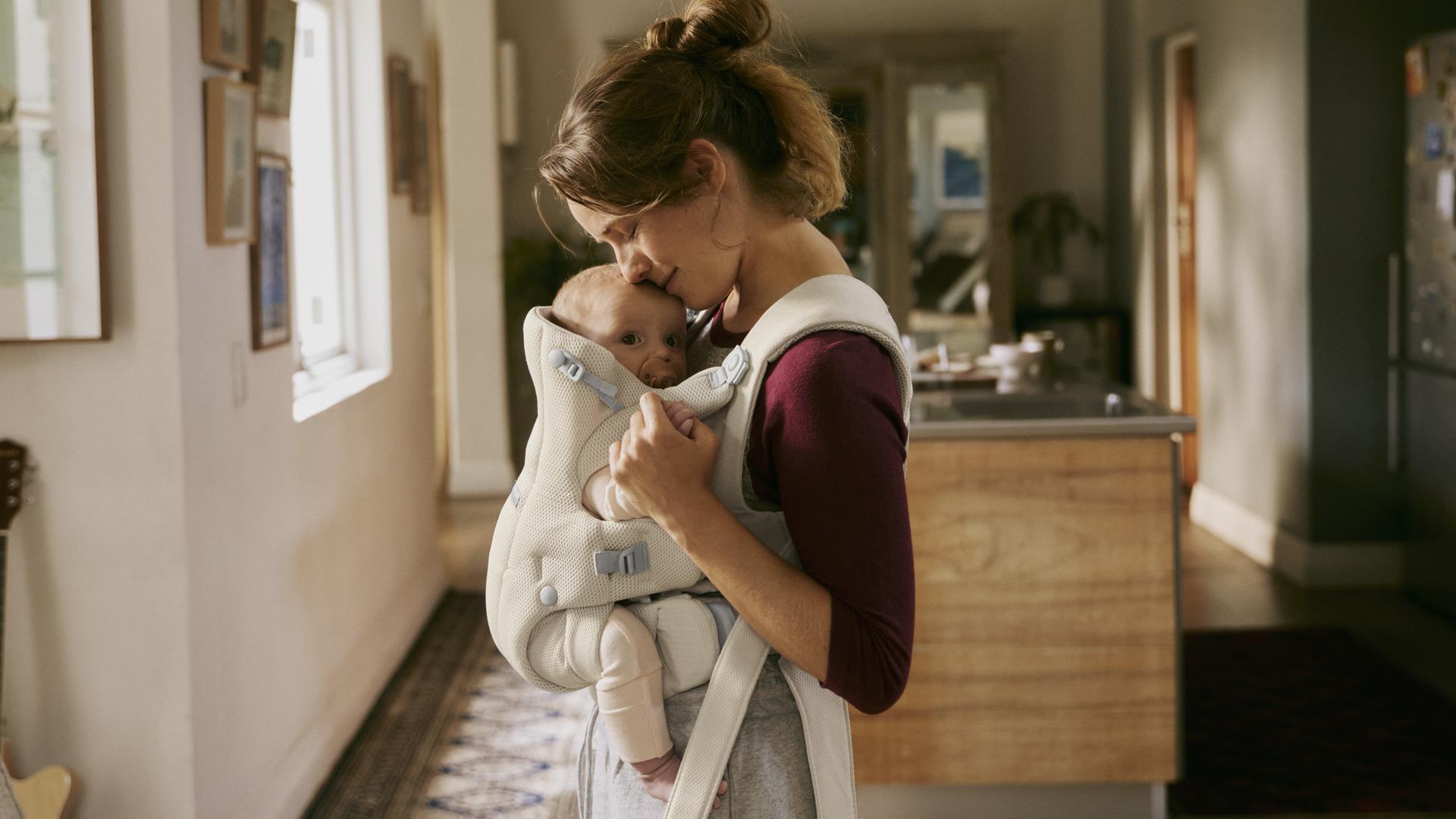 A woman tenderly kisses a baby carried in a soft white baby carrier inside a warmly lit home.