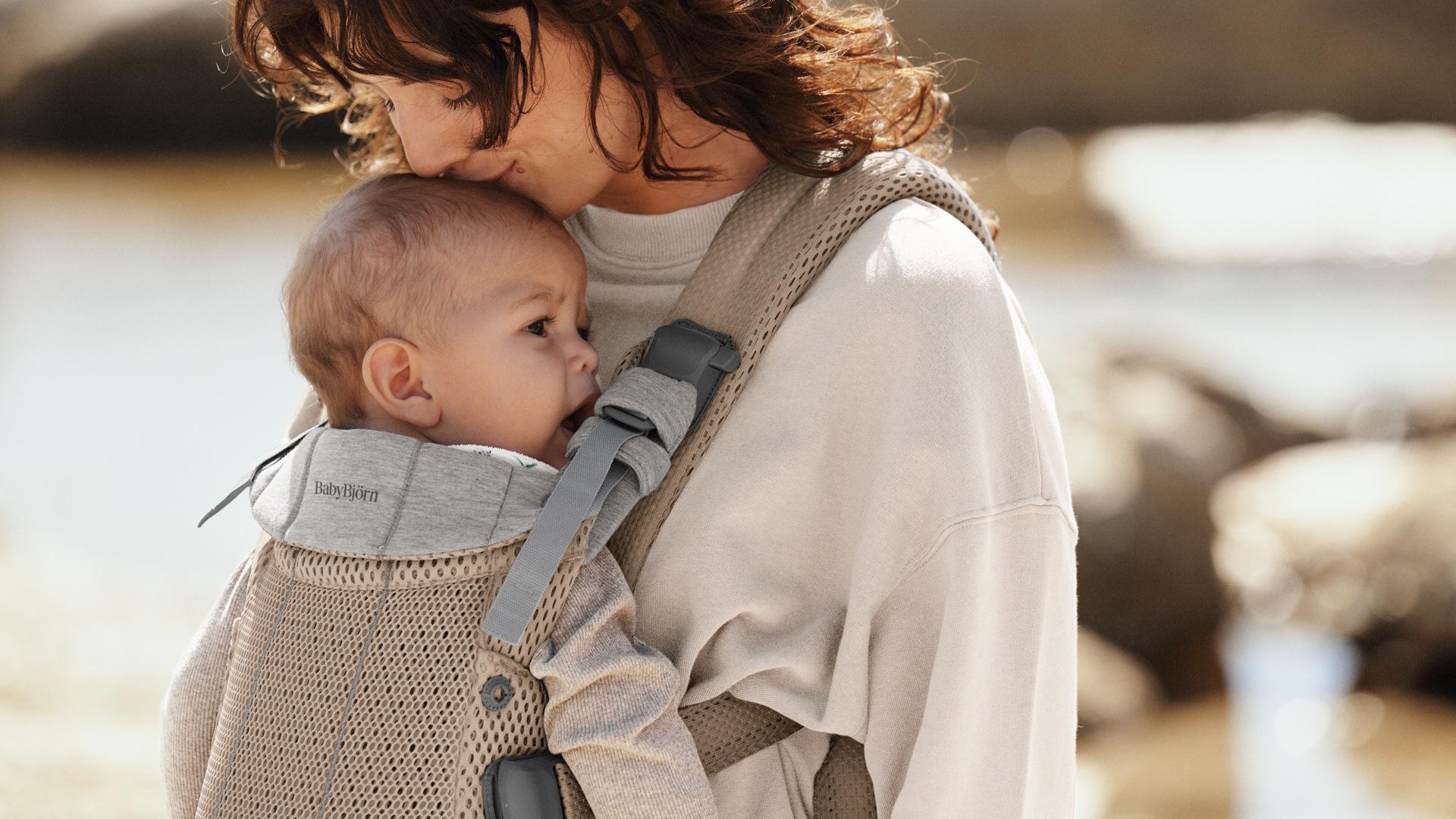 Mother gently holding her baby in a carrier, standing outdoors by a body of water, both wearing cozy clothing.