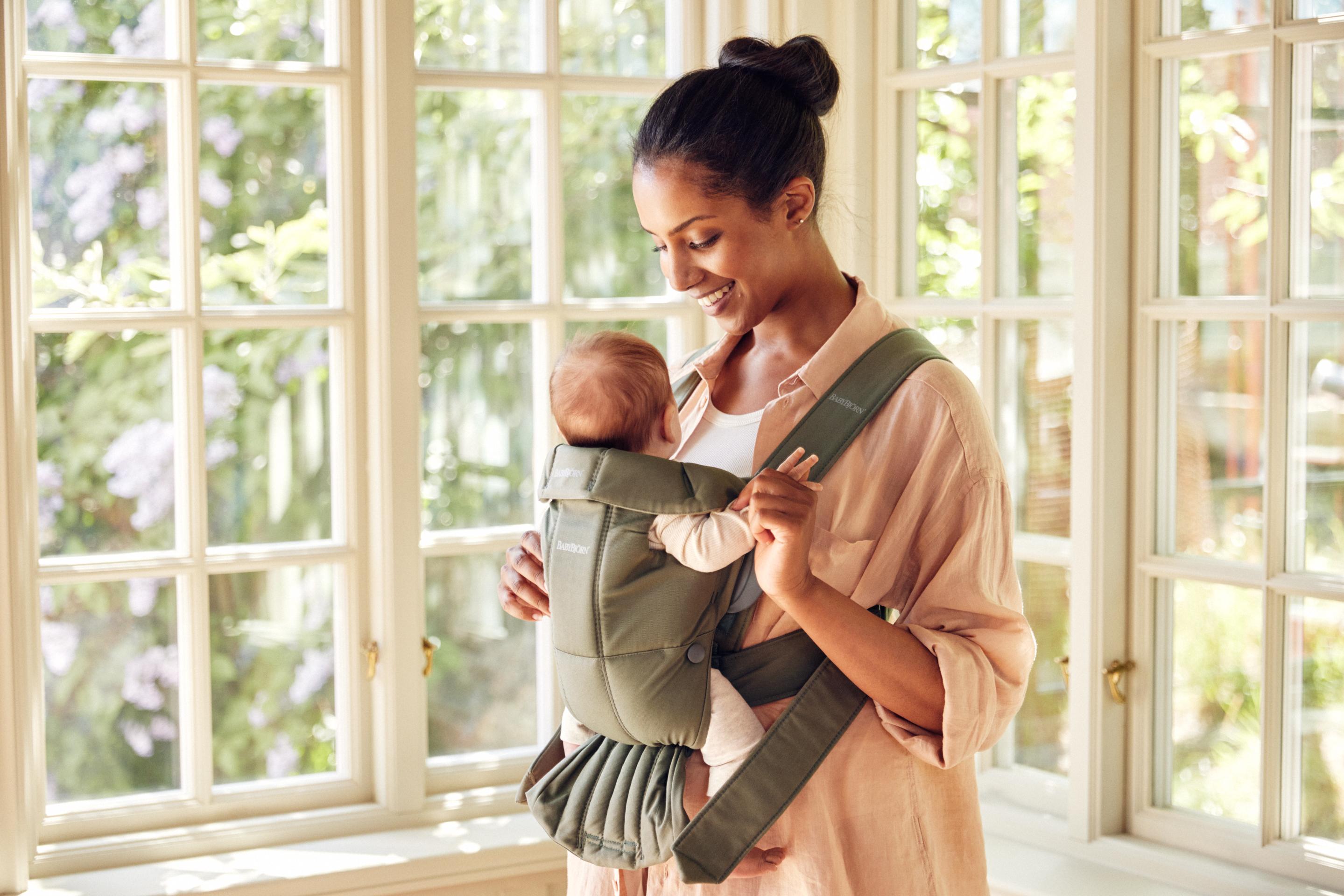 A woman stands near large windows, smiling at a baby in a green baby carrier mini from BabyBjörn. Sunlight streams in, highlighting their joyful interaction.