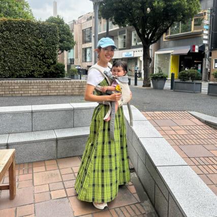 A woman in a blue cap and green plaid skirt smiles while holding a baby in a carrier. They are outdoors in a city setting with trees and buildings.
