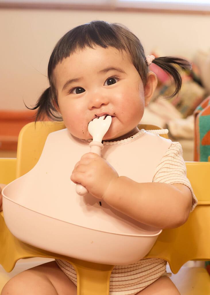 幼児がハイチェアに座り、ピンクのビブをつけてフォークを口に運んでいる。髪はツインテールに結ばれている。